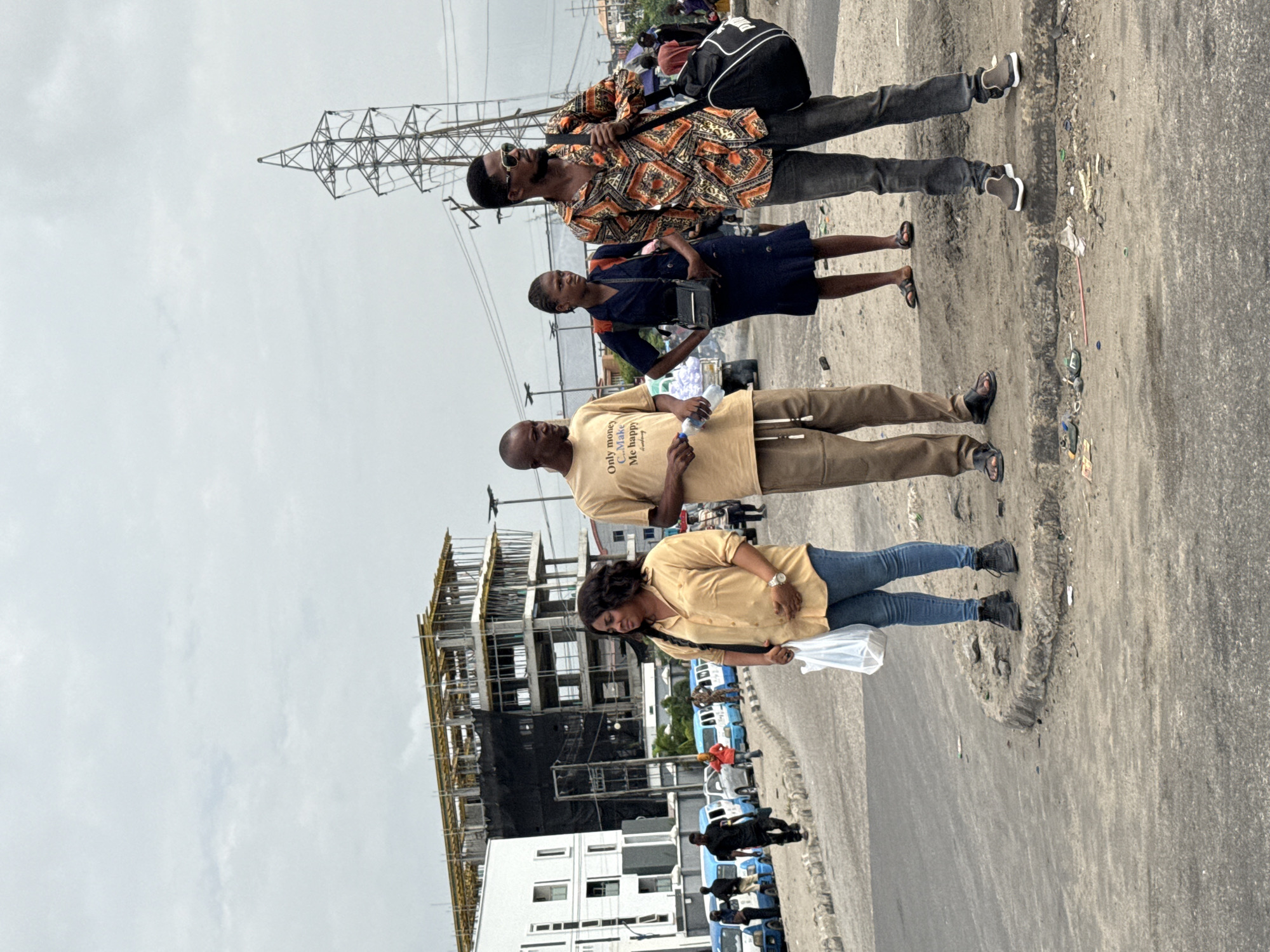 Four people are standing on a paved surface next to a road. The first person is wearing an orange patterned shirt and sunglasses and has a black bag slung over their shoulder. The second person is wearing a dark blue dress and holding a small purse. The third person is wearing a light brown t-shirt and khaki pants. The fourth person is wearing a yellow shirt and blue jeans and holding a plastic bag. In the background, there is a building under construction, power lines, and a row of blue vans. The location is Ilado, Nigeria.