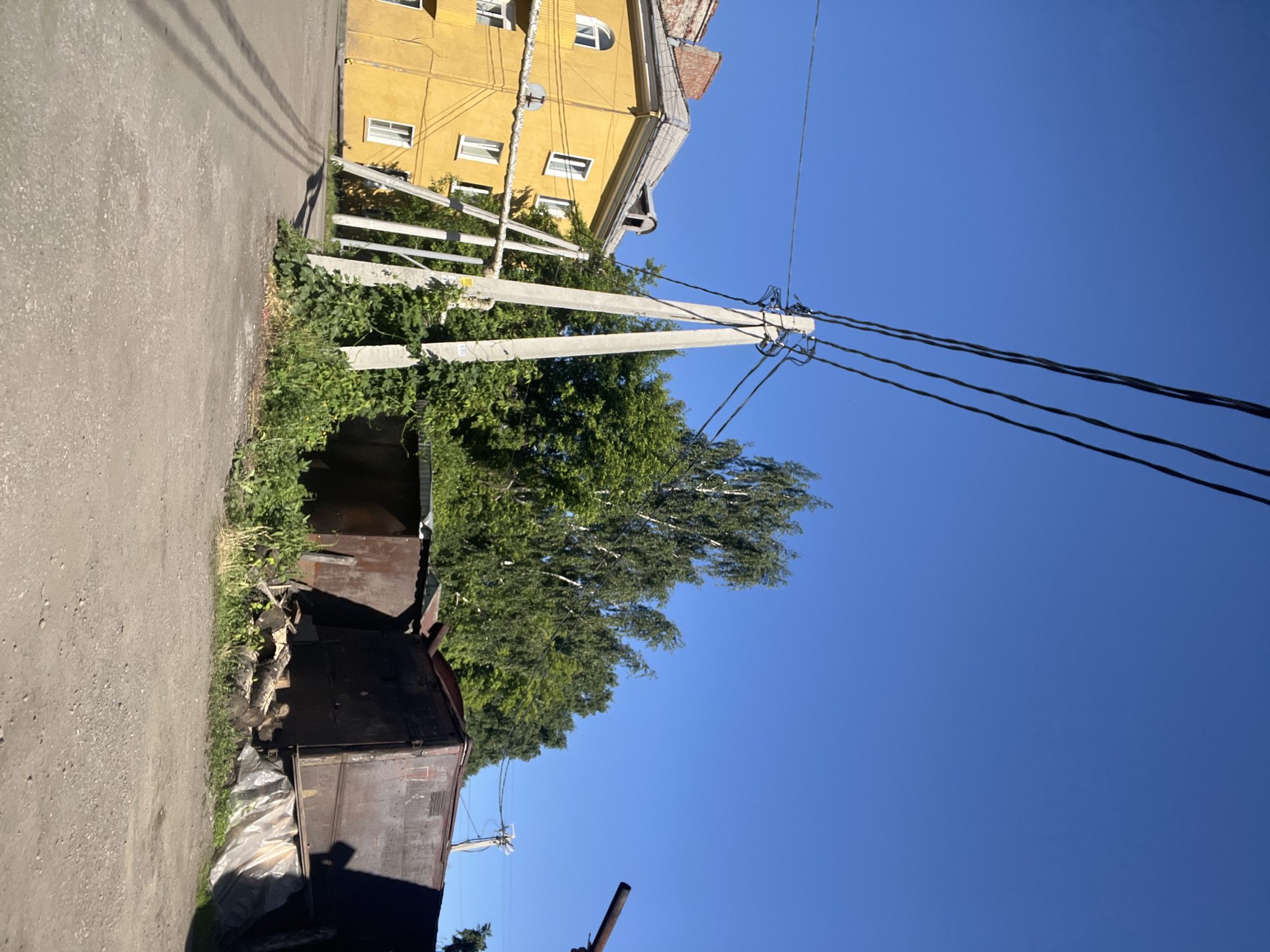 This image depicts an outdoor scene, likely in Kiselyovsk, Russia. The orientation of the image is rotated 90 degrees counter-clockwise. In the foreground, a gravelly path runs along the left side of the frame, bordered by overgrown greenery and what appear to be rusty metal sheds or structures. A large tree with green foliage dominates the center-right of the image, its branches reaching upwards.
Behind the sheds and greenery, a bright yellow building stands prominently. It has multiple windows, some rectangular and some arched. The building appears to be multi-story, with a pitched roof. Electrical poles and numerous wires are visible throughout the image, crossing the frame and connecting to the yellow building and other unseen structures. The sky is a clear, vibrant blue, suggesting fair weather and a time of day when the sun is high, likely midday or early afternoon, casting distinct shadows from the overhead wires.
There is no discernible activity or event taking place; the scene appears to be a static view of an urban or semi-urban environment. Notable details include the juxtaposition of the old, weathered sheds with the more well-maintained yellow building, and the extensive network of power lines. The overall impression is one of a somewhat unkempt but functional area under a bright, clear sky. No text is visible in the image.