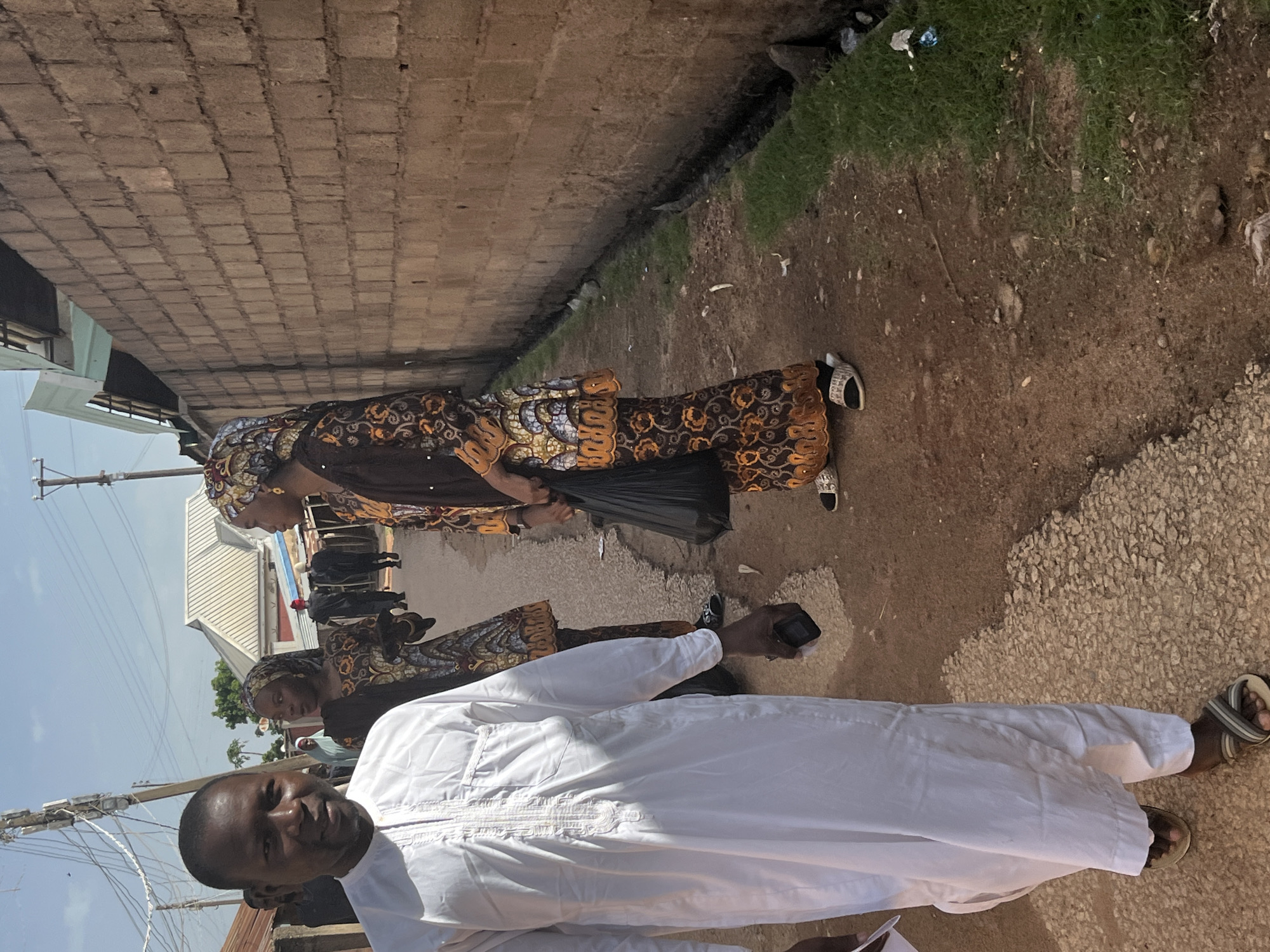 This is a vertical, eye-level, medium shot of a group of people outdoors in what appears to be a somewhat dusty, unpaved area. A man in the foreground is wearing a white tunic and is looking directly at the camera with a slight smile. Behind him and to his left, a woman in a brown and orange patterned outfit and headscarf is visible, holding a black plastic bag. Further back, two more individuals in similar patterned attire are seen. The ground is a mixture of dirt, gravel, and some patches of green grass. A rough brick wall runs along the left side of the frame, and a less defined wall or structure is visible on the right. In the background, there are glimpses of buildings with metal roofs. The lighting suggests it is daytime, possibly mid-morning or afternoon, with a bright sky and shadows. The overall scene conveys a sense of everyday life or a community gathering. No discernible text is present in the image. The context provided indicates the location is Jos, Nigeria.