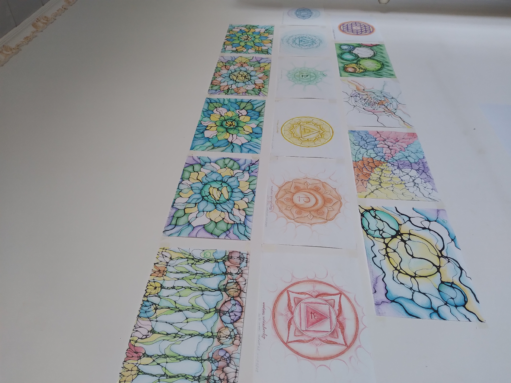 A wall in a room displays numerous pieces of artwork. The artwork is arranged in horizontal rows. The top row features abstract, colorful designs resembling flowers or mandalas, created with thin black lines and watercolor paints. The second row primarily consists of circular, mandala-like drawings, some with vibrant colors like orange and yellow, while others are in shades of green and blue. The bottom row exhibits more abstract, fluid designs with a mix of colored lines and shapes, some resembling neural networks or organic patterns. The wall itself is white, and the lighting suggests an indoor environment. No people or specific city locations are visible in the image. The artwork appears to be framed or mounted on paper.