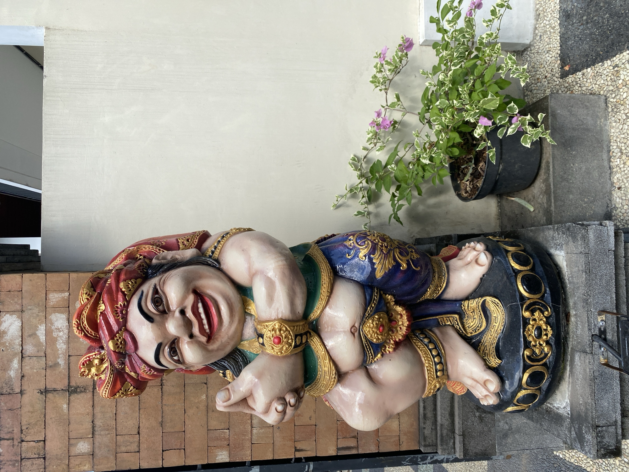 The image depicts a colorful statue of a reclining, smiling figure with a red and gold head covering, positioned near a wall and a potted plant. The statue has gold and blue detailing on its clothing and is lying on a black base with gold accents. The location is North Kuta, Indonesia. The statue appears to be outdoors, near a stone-paved area and a brick wall, with a building visible in the background. The potted plant next to the statue has green and white foliage with small purple flowers.