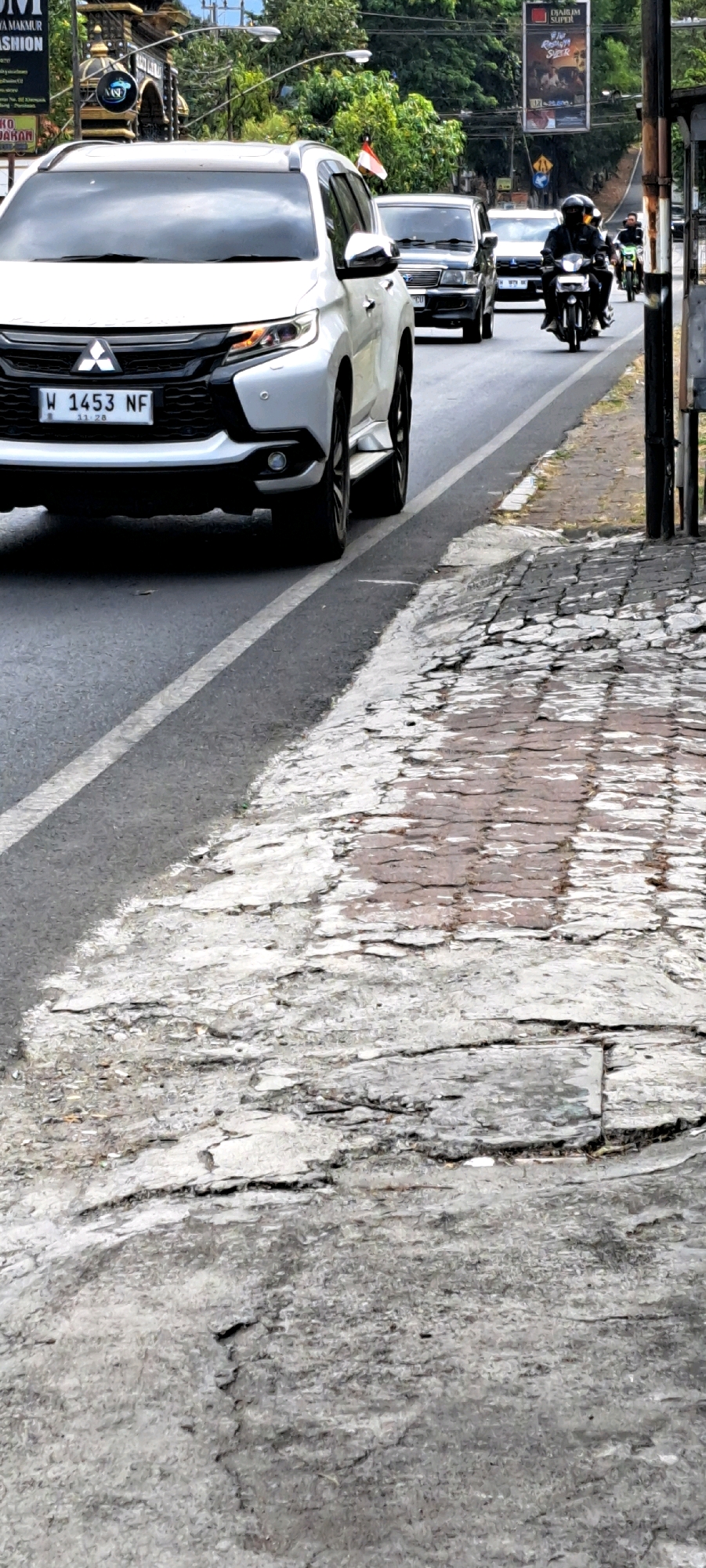 This is a medium-distance, eye-level shot of a street scene in Pandaan, Indonesia. The foreground features a cracked and worn cobblestone sidewalk adjacent to a paved road. A white SUV, identified by a license plate reading "W 1453 NF", is prominently parked on the right side of the road, facing away from the viewer. Behind the SUV, two cars and two motorcycles are visible on the road, moving in the same direction. The motorcycle riders are wearing helmets. In the background, trees and various signs, including an advertisement, line the street. A flagpole with an Indonesian flag is also visible. The overall scene depicts a typical day on a busy street in the city.