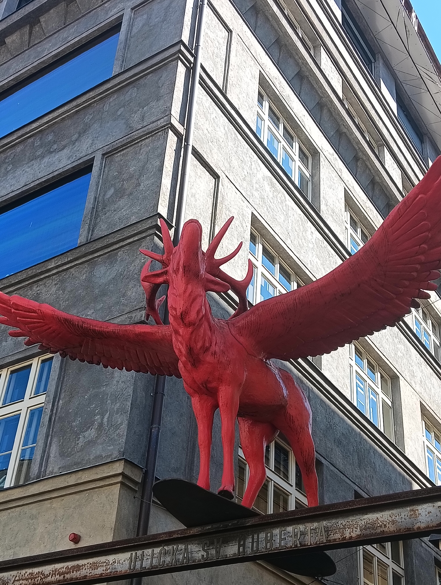 An upward-angle, medium shot captures a bright red sculpture of a winged deer mounted on a weathered metal beam against a textured, grey concrete building in Prague, Czechia. The building features rows of rectangular windows, some with blue reflective glass and others with white panes. The sculpture, detailed with a stag's head, antlers, and feathered wings, is the central focus. The metal beam below the sculpture has embossed letters that are partially obscured by rust and wear. The scene is outdoors, with a clear, bright sky visible in the upper right portion of the frame.