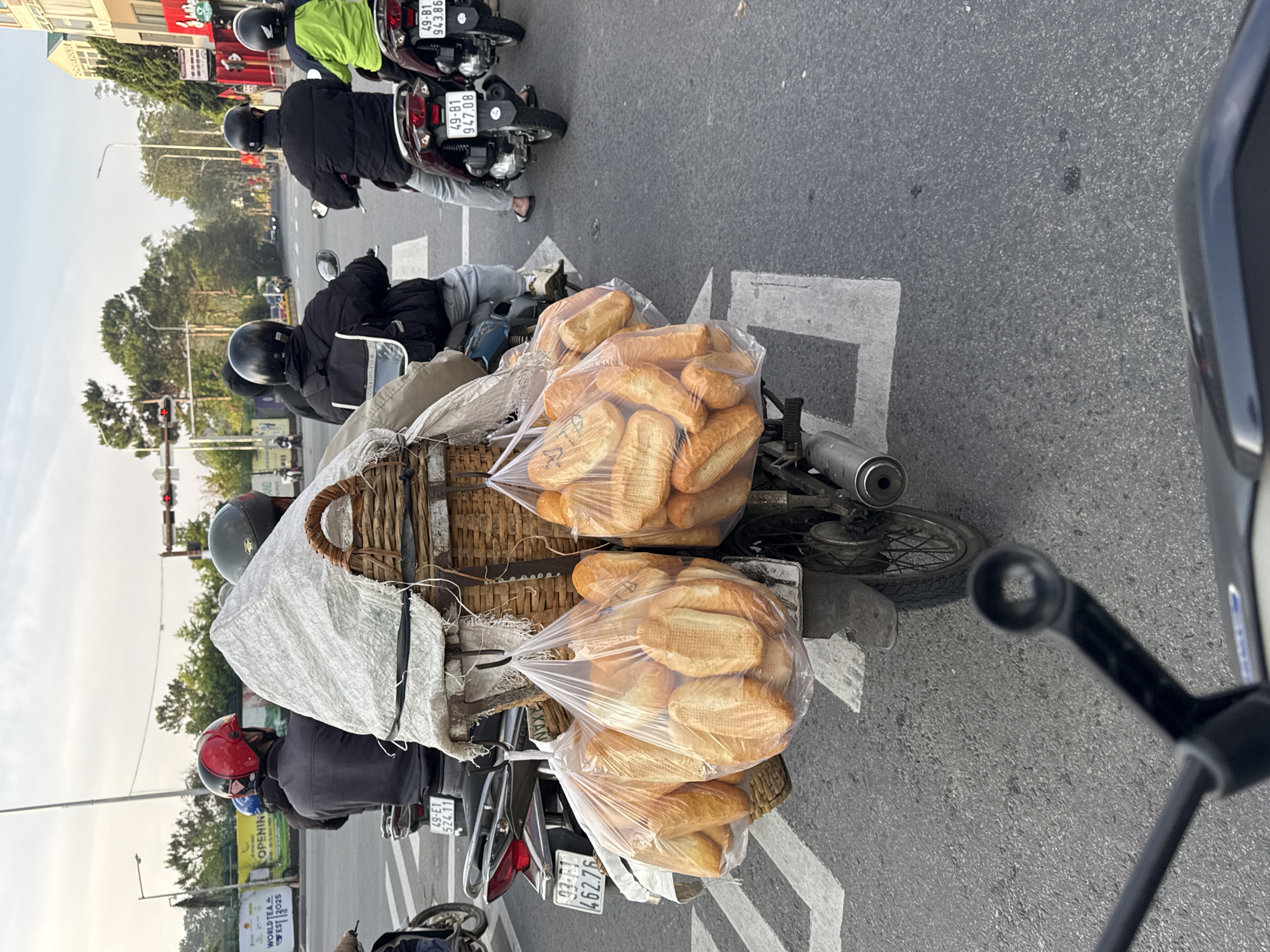 This street-level image captures a bustling scene in Da Lat, Vietnam, featuring several people on motorcycles, one heavily laden with bread. The primary subject is a motorcycle in the foreground, equipped with large wicker baskets on its back, overflowing with multiple clear plastic bags containing freshly baked baguettes (bánh mì). The rider, wearing a red helmet and a dark jacket, is part of a line of motorcycles stopped in traffic.
Other individuals on motorcycles are visible, all wearing helmets – some black, one light green. They appear to be waiting at a junction, indicated by traffic lights in the distance showing red, and white road markings on the asphalt. The setting is an urban environment, with buildings and lush trees lining the street in the background under a bright, overcast sky, suggesting daytime.
The image is captured from the perspective of another motorcycle rider, with a portion of a handlebar visible in the extreme right foreground. The scene depicts a common sight of daily commerce and transportation in Vietnam, with bread being delivered or sold.
Visible text includes several motorcycle license plates, such as "49-E1 524.11," "49-81 947.08," and "49-81 943.86," which are prefixes common to Lâm Đồng province where Da Lat is located. A banner in the background features text like "WORLD TEA," "EST 2025," and "OPENING."