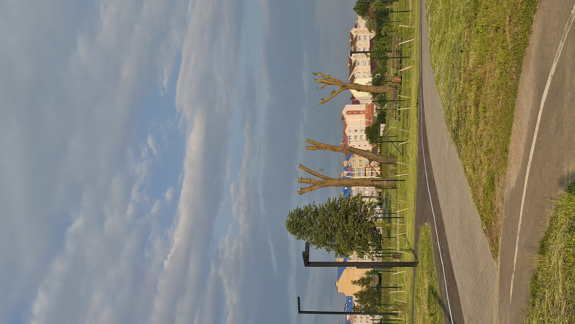 The image depicts a park area with a paved pathway in the foreground, curving to the right. The pathway is bordered by grass on both sides, with some of the grass appearing recently mowed. In the middle ground, there are three trees with their branches pruned back, appearing bare and stylized. To the left of these pruned trees, a single, full-foliaged tree stands. Beyond the trees, a row of multi-story residential buildings with colorful facades and blue roofs are visible. The buildings are of modern design, suggesting a residential development. The sky above is partly cloudy, with patches of blue sky interspersed with grey and white clouds. Two modern-style black lampposts are also present in the middle ground, one on the left and another to the left of the pruned trees. The scene appears to be set in an urban or suburban environment, likely a public park or green space. This park is located in the city of Smaliavichy, Belarus.