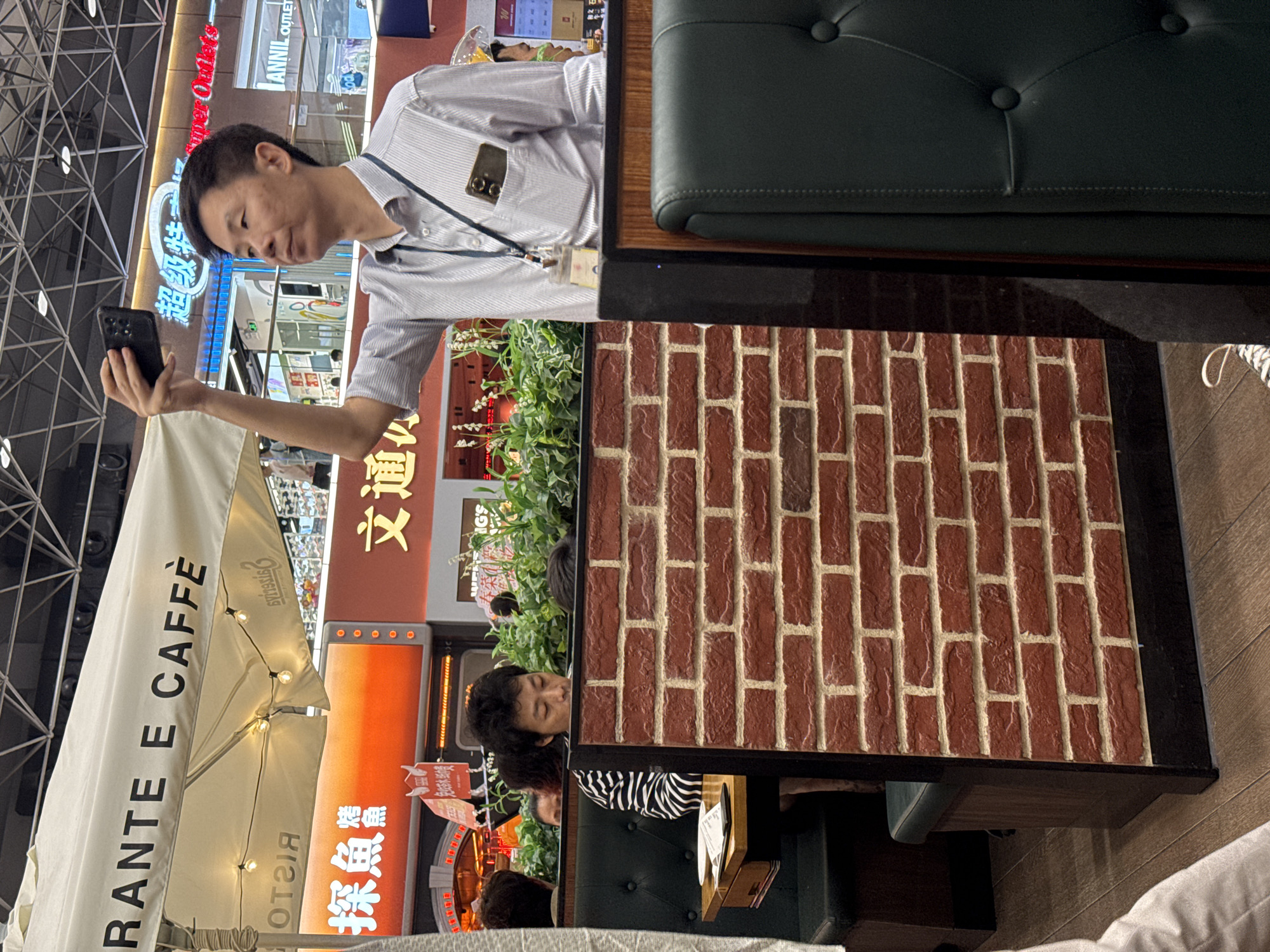The image depicts a man in Haizhu District, China, holding a smartphone while sitting at a table outside a restaurant named "RANTE E CAFFÈ". He is wearing a light-colored shirt with a lanyard and ID badge. The table is adorned with a brick pattern and faces the street, where storefronts display Chinese characters and English signage. The location appears to be a shopping area or outdoor mall. A green cushioned booth seat is adjacent to the table. There are other people dining at tables in the background, including a person in a striped shirt. An awning partially covers the outdoor seating.