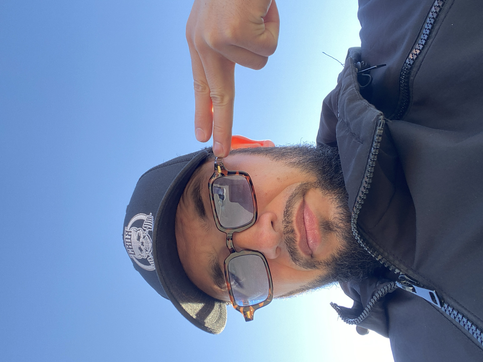 The image shows a close-up, low-angle selfie of a man with a beard, wearing a black baseball cap with a skull logo and a black jacket with a zipped collar. He is also wearing tortoiseshell-patterned glasses. His right hand is raised, with two fingers extended towards his right temple, near his glasses. The background is a clear, bright blue sky, suggesting an outdoor setting. The man's head is tilted slightly upwards and to his left. The image is shot from a perspective looking up at the man. There is no discernible indication of the city of Koskolovo, Russia, or any other specific location within it.