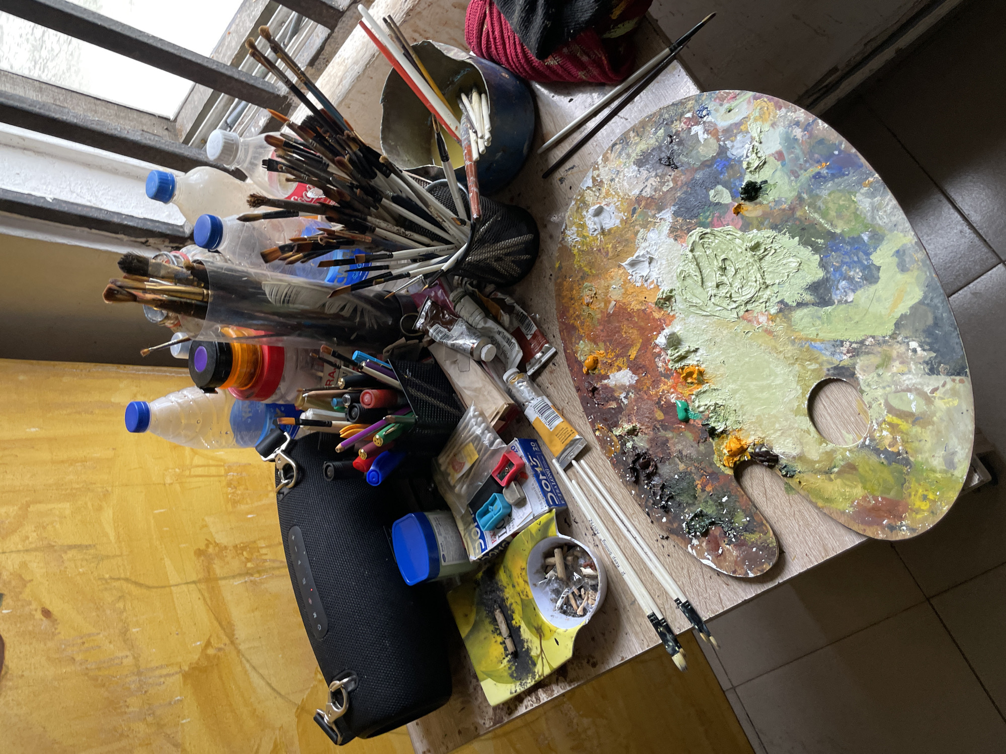 The image depicts an artist's workspace, likely in Ado, Nigeria. A well-used wooden artist's palette, covered in a variety of oil paints in shades of green, yellow, orange, red, blue, and brown, dominates the foreground. Several paintbrushes are scattered around the palette, with two longer brushes resting on top of it.
In the middle ground, various art supplies are organized on a wooden surface. A black portable speaker is positioned to the left. To its right, a collection of paintbrushes is housed in a black mesh container. Several plastic water bottles with blue caps are visible, alongside tubes of paint and other small art supplies like pencil sharpeners and cotton swabs. A ceramic bowl filled with cigarette butts and ash sits on a yellow object, possibly a palette tray.
The background shows a textured yellow wall and a window with metal bars, suggesting an indoor setting. The overall impression is one of a creative, albeit somewhat cluttered, environment where artistic endeavors are actively taking place. The presence of cigarette butts and ash might indicate a moment of pause or contemplation during the artistic process. The lighting suggests it is daytime, with natural light filtering through the window.