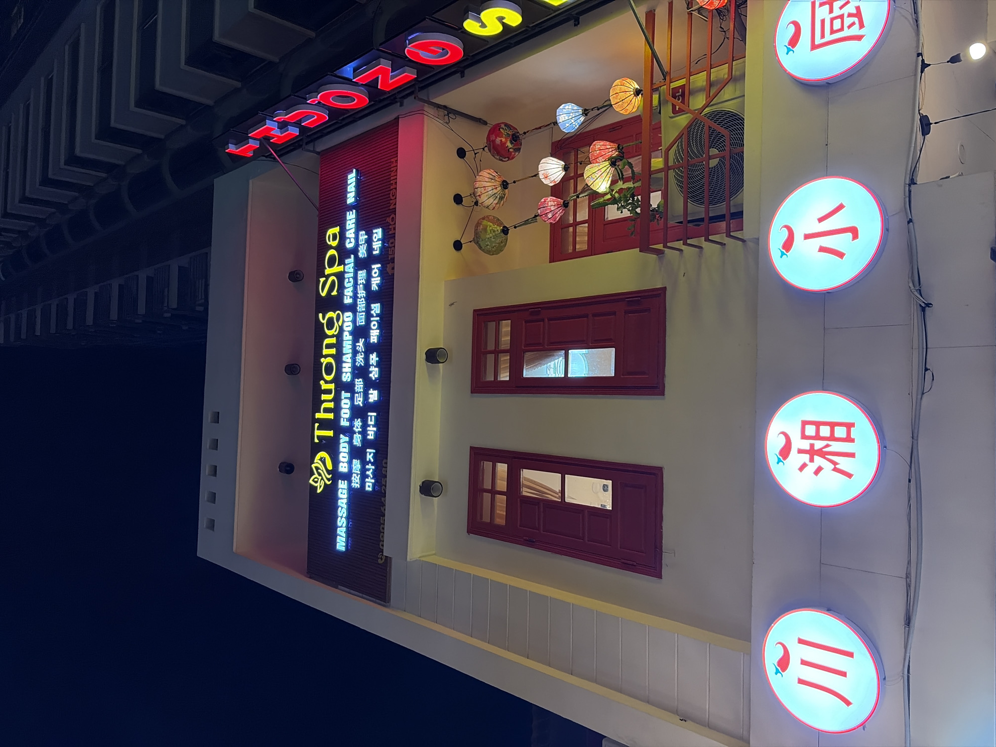 This is a medium-angle, night-time, exterior shot of a multi-story building in Da Nang, Vietnam, featuring a spa and other businesses. The building's facade is illuminated by neon signs and decorative lanterns.
At the top of the building, a prominent neon sign reads "Thương Spa" in yellow and blue lettering. Below this, a smaller sign lists services in English, Chinese, and Korean, including "MASSAGE BODY FOOT SHAMPOO FACIAL CARE NAIL". A phone number, "0905 44 25 80", is also visible. To the right of the "Thương Spa" sign, a vertical neon sign spells out "TRSONG" in red and yellow.
The building has two distinct red-framed windows on the second floor. Decorative, colorful paper lanterns are strung across the facade between the second and third levels, casting a warm glow. An air conditioning unit is visible below the lanterns.
At the bottom of the building, four circular illuminated signs are mounted. From left to right, these signs feature red Chinese characters on a light blue background. The first sign displays three vertical red lines. The second sign shows the character "湘". The third sign displays the character "小". The fourth sign shows the character "扇". Each circular sign also has a small red chili pepper design at the top.
The image is taken from a street-level perspective, looking upwards. The overall impression is one of a bustling commercial street at night. The upper portion of a tall, modern building is visible in the background, silhouetted against the dark night sky.