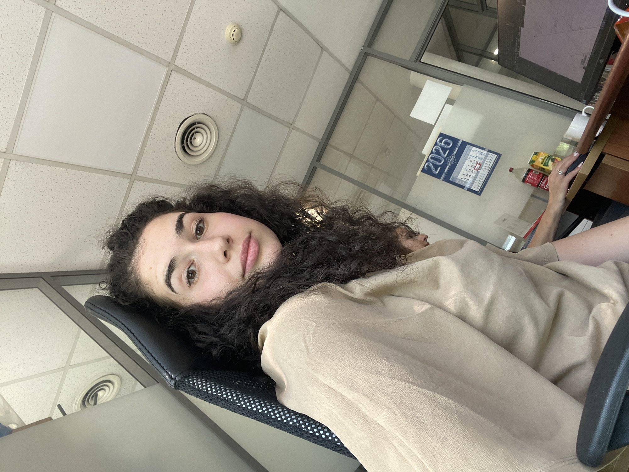 A woman with dark curly hair sits in an office chair in what appears to be an office environment. She wears a light brown or beige top. The office has a tiled ceiling with circular air vents and a fire alarm system. A glass partition is visible, separating her workspace from another. On the desk behind her, there is a calendar displaying "2026" and a mouse being held in the person's right hand. There is a juice box and a can next to the hand. The image appears to have been taken from a low angle. The location is potentially in an office building, but there is no specific context indicating that the image was taken in Sofia, Bulgaria.