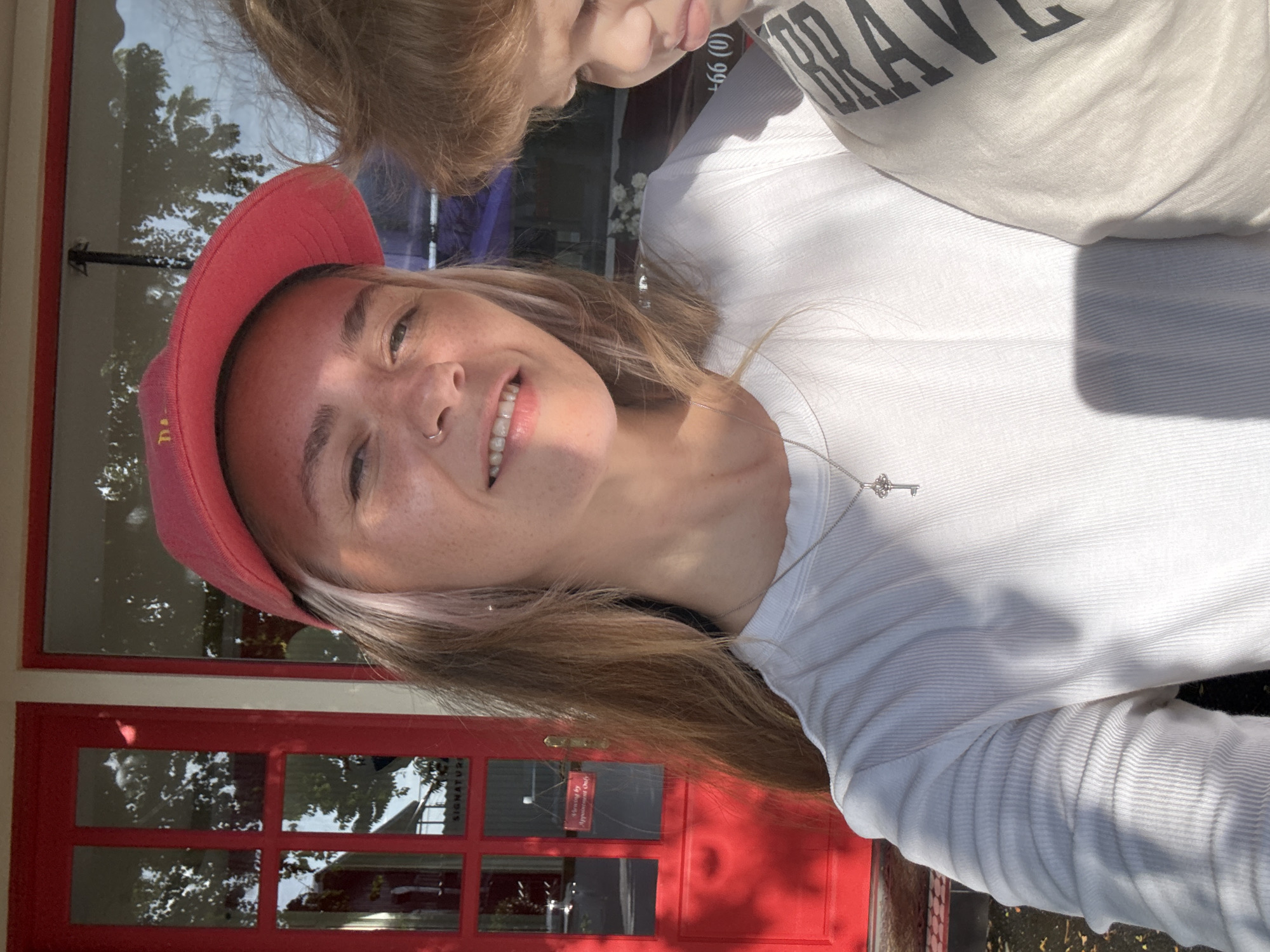 In this close-up, selfie-style photograph, a woman wearing a red baseball cap and a white ribbed long-sleeved shirt is pictured. She has blonde hair with streaks of pink, a nose ring, and is smiling. She is wearing a silver necklace with a key pendant. Next to her, a young child with blonde hair is partially visible, wearing a light-colored shirt with dark lettering. The background features a bright red door with glass panes, reflecting trees and sunlight. A sign on the door reads "Viewing by Appointment Only". The scene appears to be outdoors, possibly in front of a shop or establishment in Choeng Thale, Thailand, as suggested by the reflective glare on the glass and the style of the door. The lighting indicates it is daytime, with strong sunlight creating shadows.