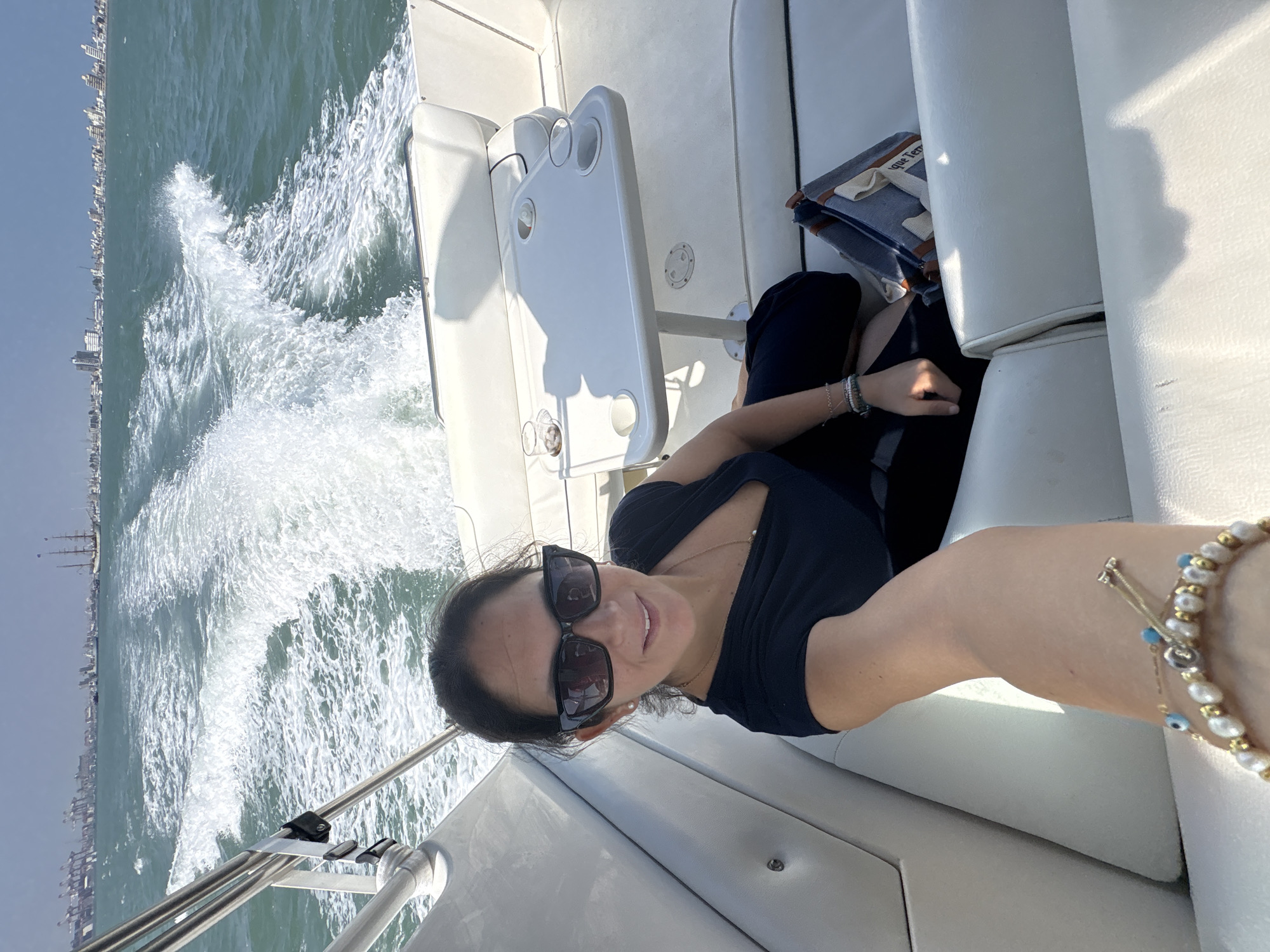 A selfie taken on a moving boat off the coast of Callao, Peru. A woman wearing sunglasses and dark clothing sits on a white boat seat, with a small table next to her. In the background, the wake of the boat churns the water, with the city skyline of Callao visible in the distance under a light blue sky. A bracelet is visible on the woman's wrist.