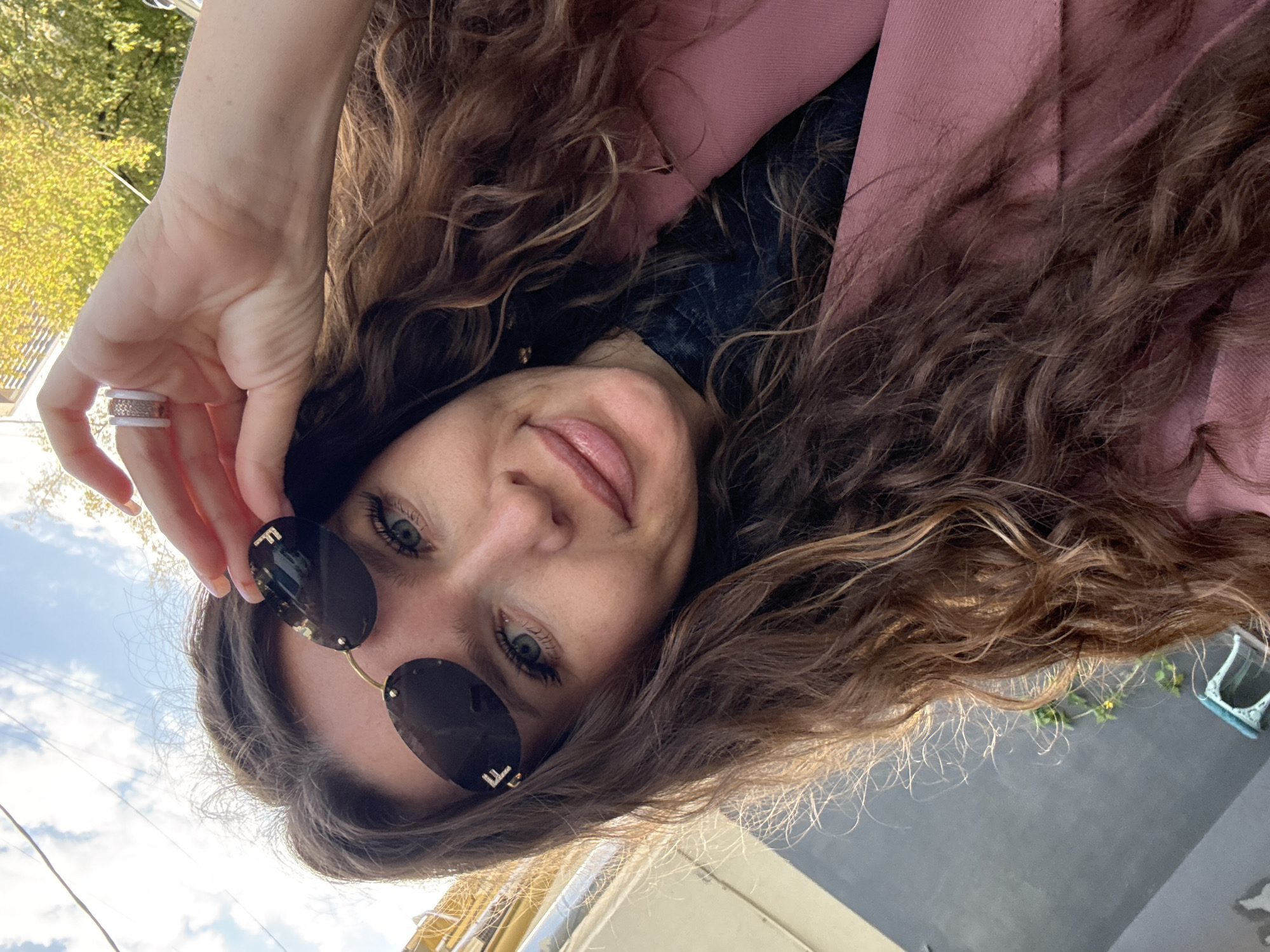 In a close-up selfie, a woman with long, curly brown hair is pictured outdoors, likely in Moscow, Russia, given the context of previous queries. She is wearing oval-shaped, rimless sunglasses with gold accents, one of which displays the "FF" logo, suggestive of Fendi. Her right hand is raised, holding the sunglasses against her forehead, revealing a textured white ring on her ring finger. She is also wearing a pink jacket over a dark top. The background features a sky with clouds, the yellow railing of a building, and foliage, indicating an urban setting. The lighting suggests it is daytime, possibly late afternoon due to the warm tones.