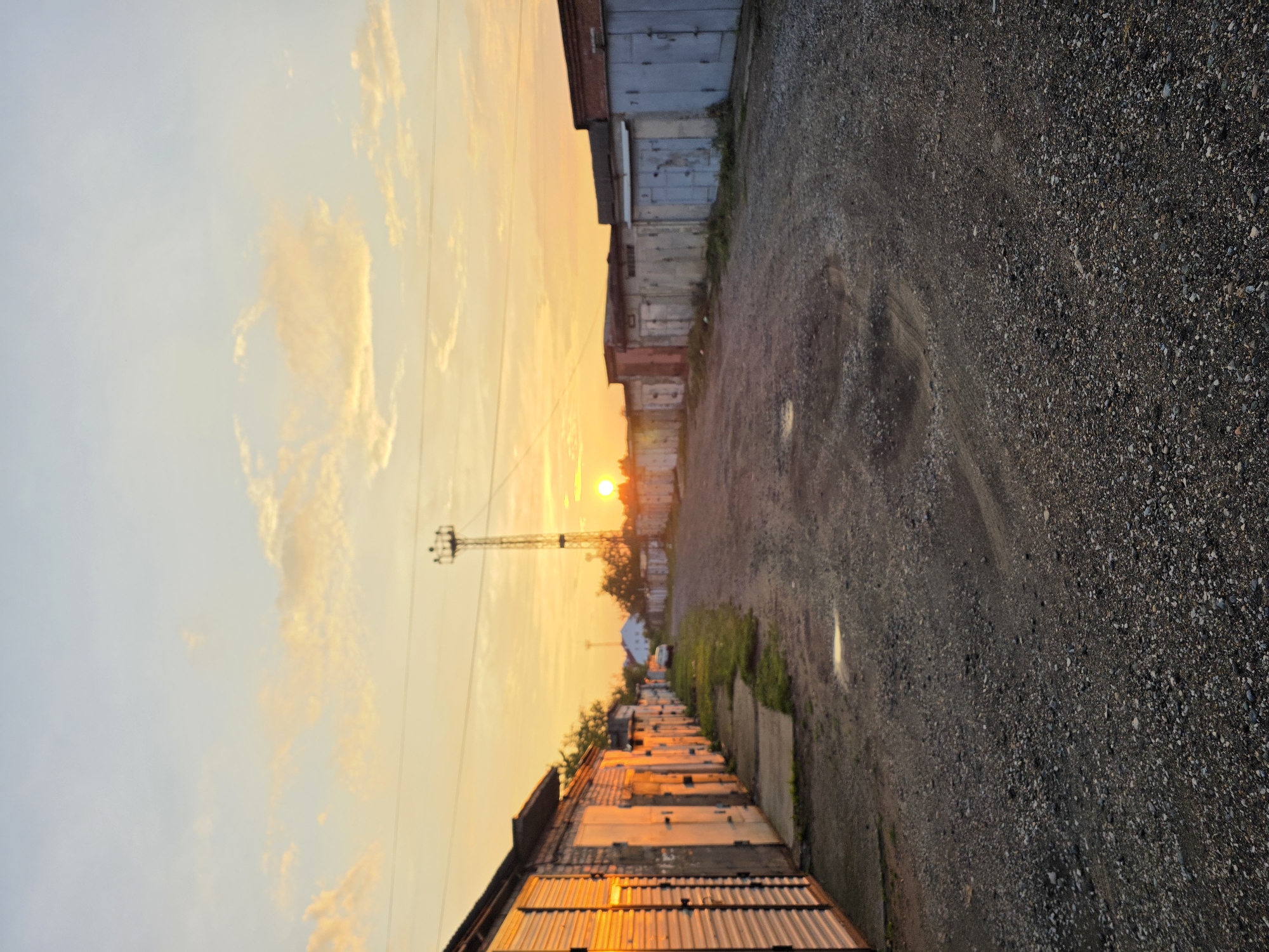 An outdoor, medium-shot, vertical photograph depicts a gravel road leading between two rows of garages at sunset in the city of Chaikovsky, Russia. The sun, a bright orange orb, is low on the horizon, casting a warm golden light across the sky and illuminating the corrugated metal siding of the garages on the left. The sky is a gradient of pale blue to orange with scattered clouds.
On the left, a line of garages with corrugated metal doors and brick construction is visible. On the right, a similar line of garages is present, constructed from concrete blocks and metal. A tall metal tower, possibly a communication tower, stands in the distance, with the sun setting behind it. A single white car is parked on the grass verge between the garages in the distance. The road is covered in gravel and shows signs of wear, with puddles reflecting the light. The scene is quiet and appears to be in a garage complex or industrial area.