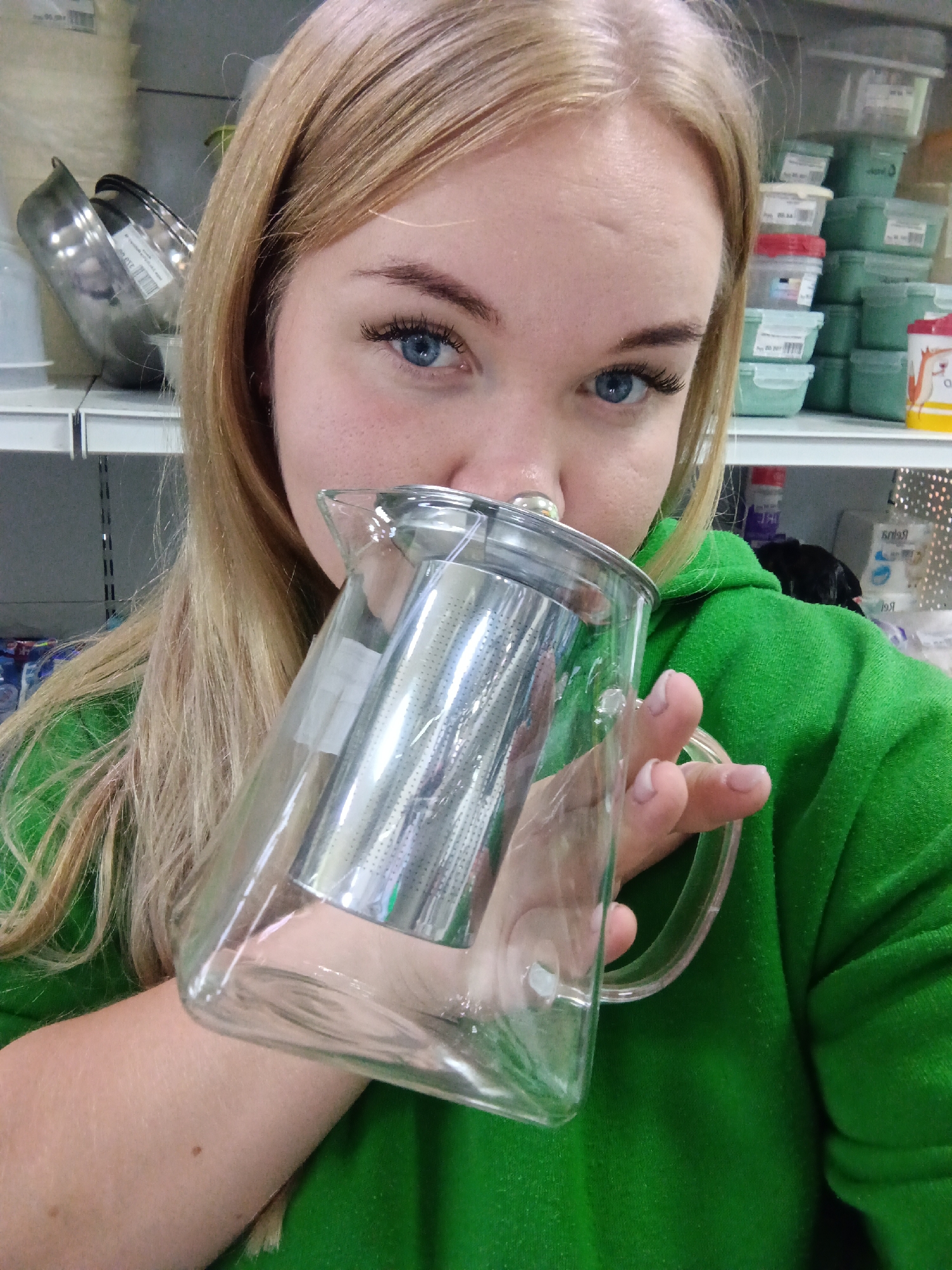A young woman is captured in a close-up, eye-level portrait, holding a clear glass teapot with a cylindrical stainless steel infuser. She looks directly at the camera with bright blue eyes, framed by long blonde hair and defined eyebrows. Her expression is neutral and engaging. She wears a vibrant green hooded sweatshirt.
The background indicates an indoor retail or storage setting, featuring white shelving stocked with various items. To the left, several silver-colored bowls or containers are visible on a shelf, one of which has a price label reading "200 РУБ" (200 Rubles). To the right, numerous light green plastic containers with lids are stacked, many bearing white labels, some clearly displaying the number "400" and barcodes. A red-lidded container within the stack also shows "400". A red and white cup with a cartoon character is partially visible among the items. Further in the background, reversed Cyrillic text, possibly "Белояр", is reflected on a shiny surface.
The woman's skin is fair, and her eyelashes are prominent. Her fingernails are short and unpolished. A small mole is visible on her right forearm. The lighting is bright and artificial, characteristic of an indoor commercial space, creating reflections on the glass teapot and metal infuser. The setting is likely a store in Фоки, Russia.