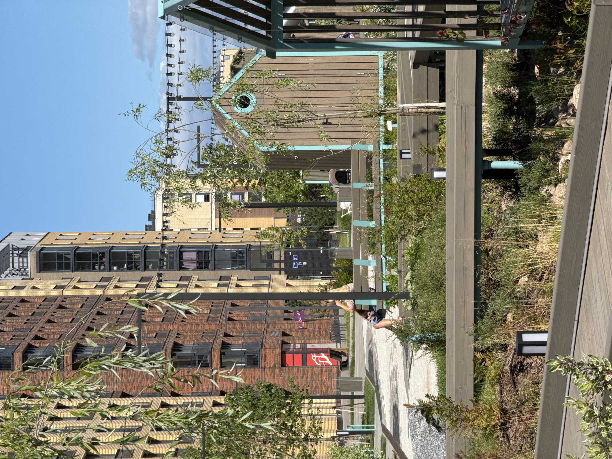 The image captures a sunny day in Novoselye, Russia, showcasing a modern urban landscape with a focus on green spaces and contemporary architecture. In the foreground, a woman with blonde hair is seated on a light blue and wooden bench, facing away from the camera. She is wearing a light-colored top and patterned shorts. The bench is part of an outdoor seating area with wooden decking and planters.
Behind the woman, a small, wooden shed-like structure with a teal trim and a round window is visible. Further back, a tall, multi-story residential building with a brick facade, predominantly in shades of red and yellow, dominates the background. Several other similar buildings are also present, indicating a newly developed residential area.
The environment is characterized by lush greenery, including trees with drooping branches and various shrubs. A pathway with a patterned surface leads into the scene, and decorative string lights are suspended in the air above the wooden structures, suggesting an area designed for outdoor leisure and social gatherings. The sky is a clear blue with a few wispy clouds.