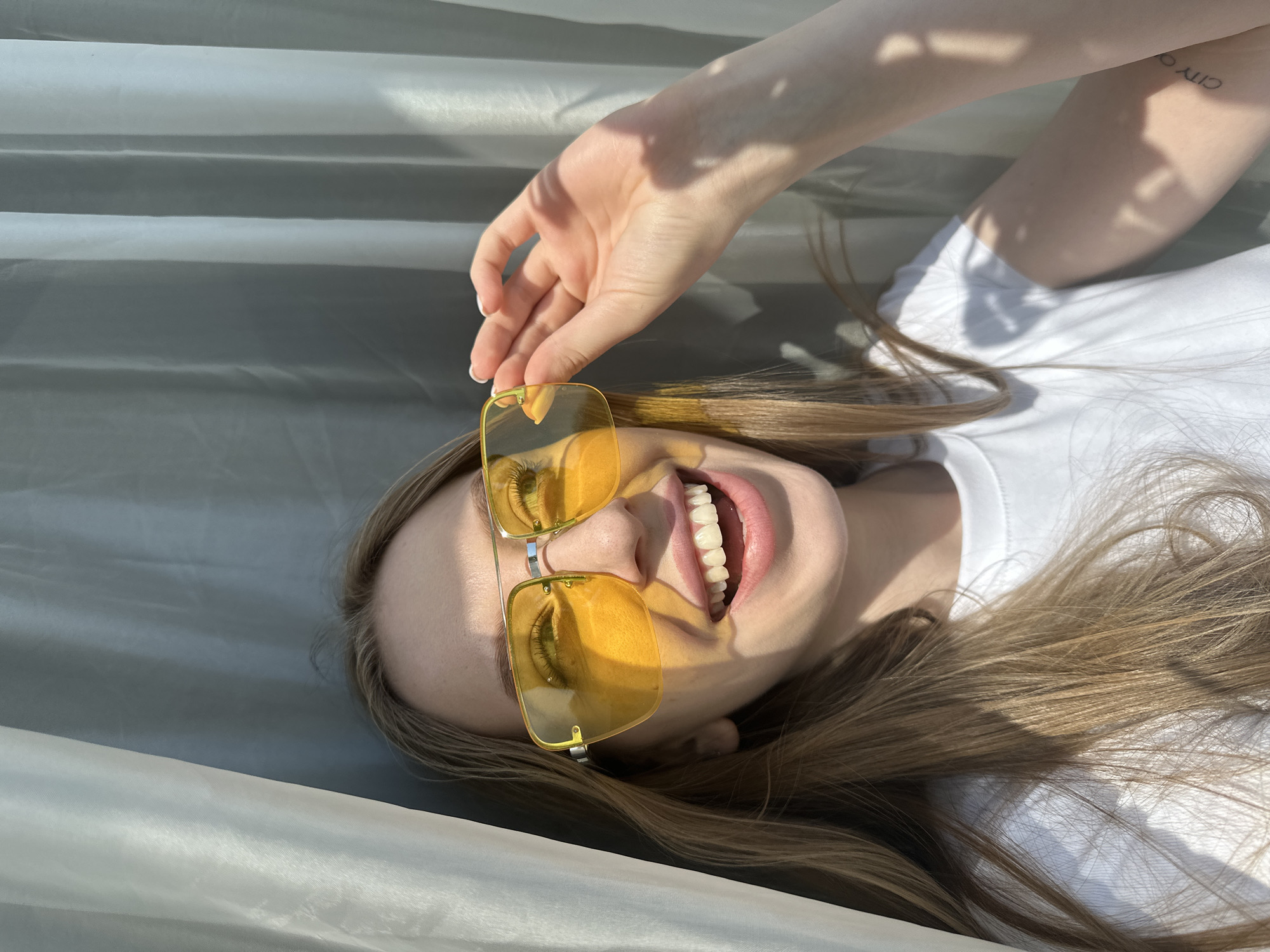 A woman wearing a white t-shirt and yellow sunglasses is lying on a gray surface, possibly fabric, with her head tilted back and her mouth open in a smile. Her left hand is touching the right lens of her sunglasses. A tattoo is visible on her right forearm. The location, Chaikovsky, Russia, cannot be confirmed from the image. The environment appears to be indoors, with sunlight casting shadows.