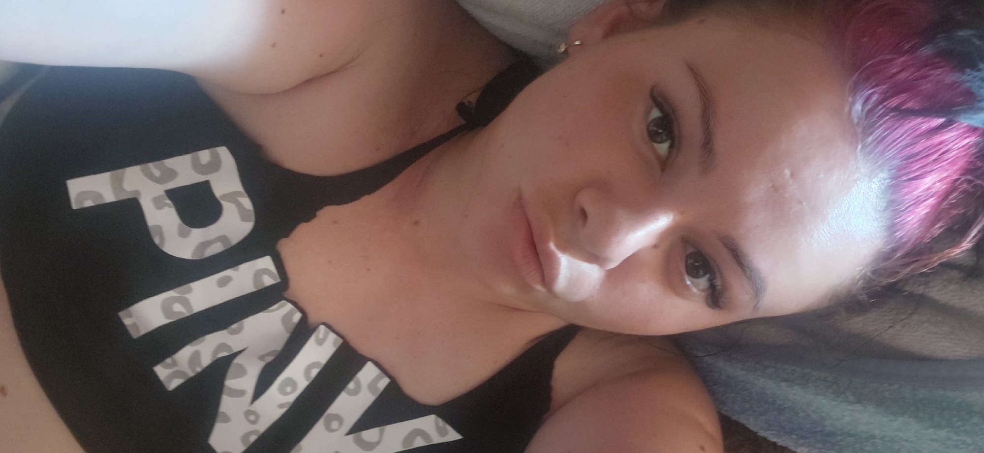 This is a close-up, overhead selfie of a young woman with brightly dyed pink hair, lying down and looking directly at the camera. She is wearing a black tank top with large, white lettering that appears to spell out "PINK," accented with a grey leopard print design. Her expression is neutral, with her lips slightly parted. The lighting suggests an indoor setting, possibly a bedroom, with a soft glow illuminating her face. The background is out of focus, showing a textured surface that could be bedding or upholstery. Her left arm and shoulder are visible, and she is wearing a small hoop earring. The overall impression is a casual, personal snapshot.