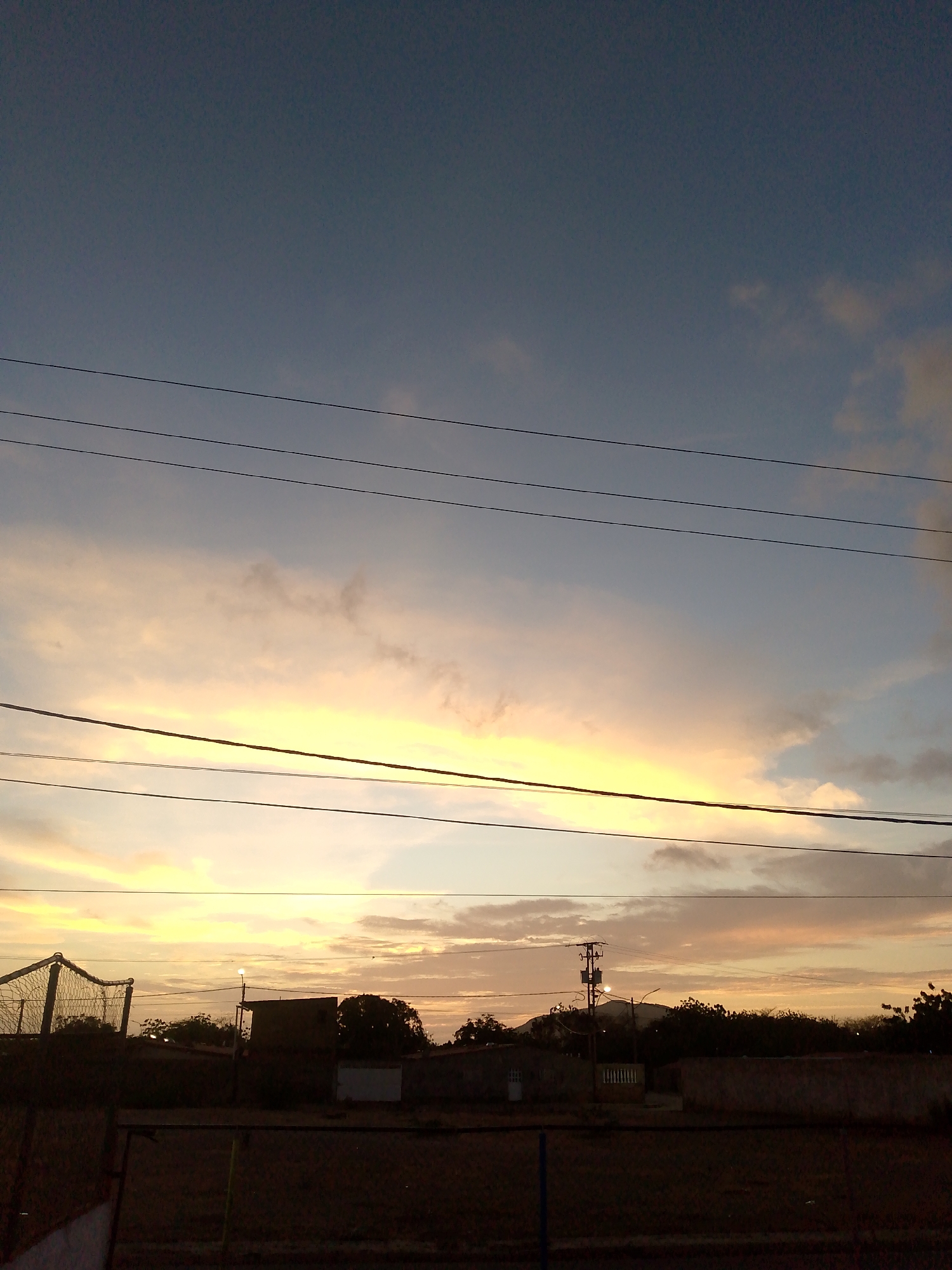 The image depicts a sunset in Altagracia, Venezuela. The sky is a gradient of dark blue at the top, fading into orange and yellow hues where the sun is setting. Several power lines stretch horizontally across the frame, obscuring the sky. In the lower half of the image, a row of buildings and trees are silhouetted against the bright sky. A fence runs along the bottom edge of the frame, suggesting the viewpoint is from a property or enclosed area. There are no people visible in the image.