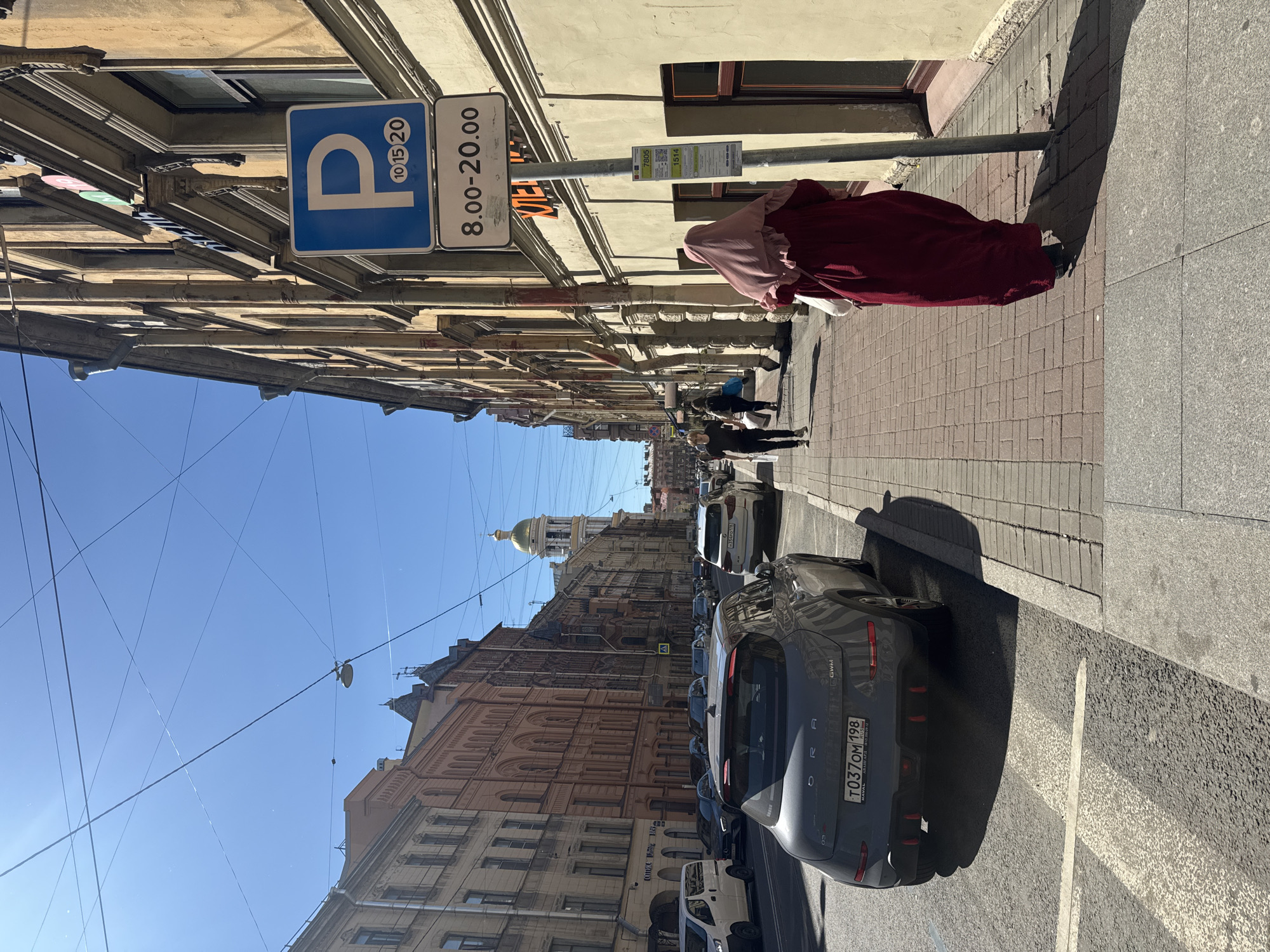 A street scene in Saint Petersburg, Russia, features a woman in a maroon hijab and dress walking on a sidewalk next to a building. She is walking away from the camera, and she is holding a white bag. A parking sign with a blue "P" and numbers 10, 15, and 20 is visible on a pole next to her. Below the "P" sign, a sign reads "8.00-20.00". Several cars are parked on the street and driving on the road. In the background, buildings line the street, and a gold-domed church is visible in the distance. The sky is clear and blue, indicating a sunny day. The sidewalk is paved with red bricks and gray concrete. The street is asphalt and has white lane markings.