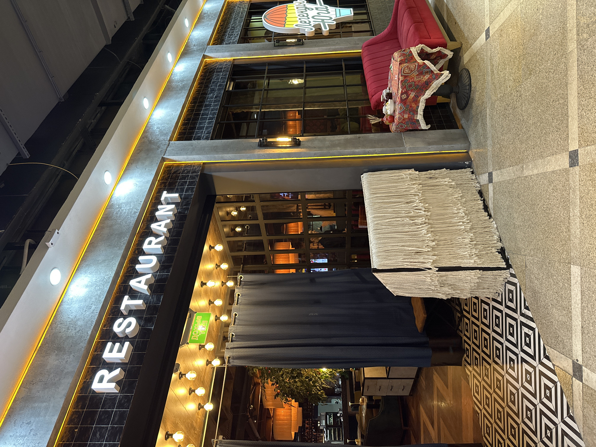 The image shows the exterior of a restaurant, identified by the large, vertically oriented "RESTAURANT" sign. The building has multiple stories. The front facade has rows of lights. Below a blue curtain is the entrance to the restaurant. A pile of papers is on the ground. On the upper floor, there is a red couch. A sign is above the red couch.
The image location is Yekaterinburg, Russia, according to the sign shown in the image.