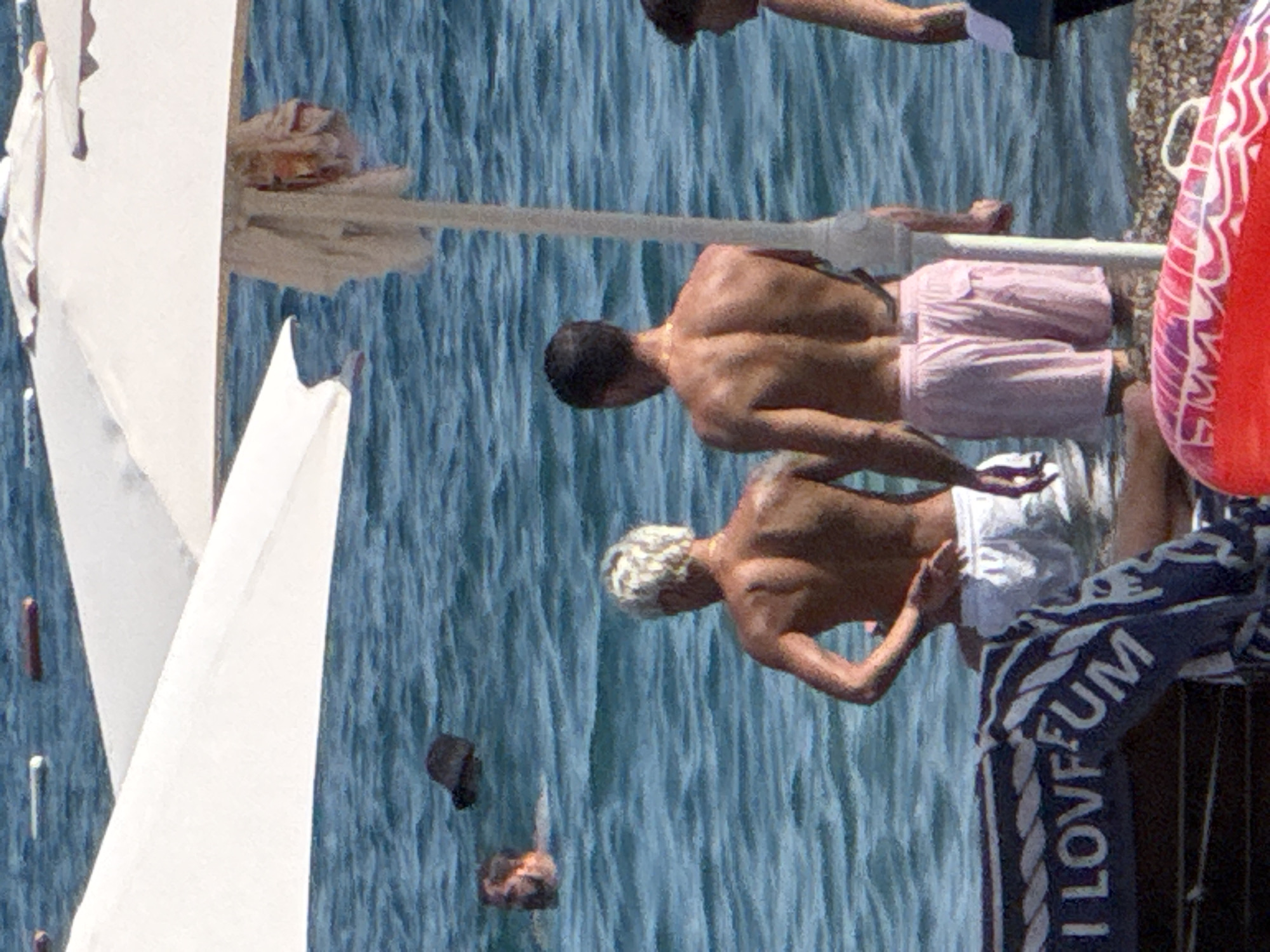The image captures a beach scene in Boreti, Montenegro. In the foreground, a person is seated on a beach chair with a towel that reads "I LOVE FUM." Behind them, three individuals are partially submerged in the blue water. Two men are facing away from the camera, one with blonde hair wearing white shorts, and the other with dark hair wearing pink swim trunks. A third person, a man with dark hair, is further out in the water, facing the shore. Two white beach umbrellas are visible, casting shadows on the water. The environment is a tranquil beach with calm waters.
