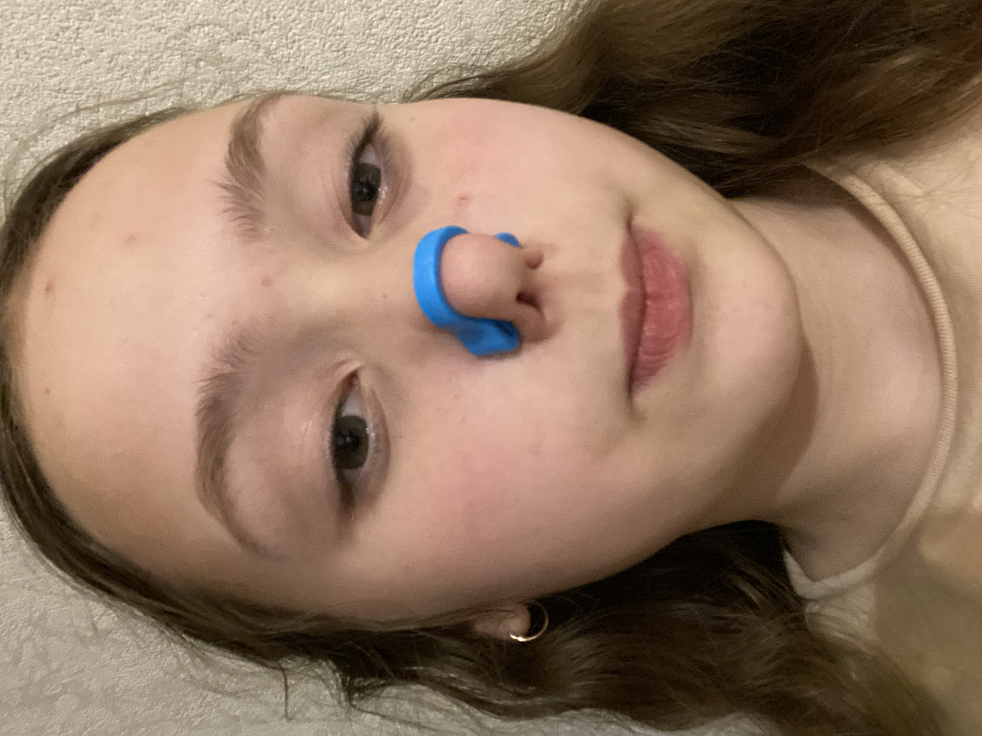 A close-up, angled portrait shows a young person, likely a girl, lying down and looking directly at the camera. She has long, dark brown hair, fair skin, and dark eyes. A prominent bright blue nose clip is affixed to her nose, covering both nostrils. Her lips are slightly parted, displaying a natural pink hue. A small gold hoop earring is visible in her left ear. Her expression is neutral and calm, with a direct gaze. She is wearing a light-colored top, possibly beige or light brown, with a visible ribbed texture around the neckline. The background is a soft, textured, light-colored surface, suggesting an indoor setting such as a bed or sofa. Small, reddish blemishes are visible on her forehead and cheek. There is no visible text. The time of day is not discernible, but the lighting is consistent with an indoor environment.