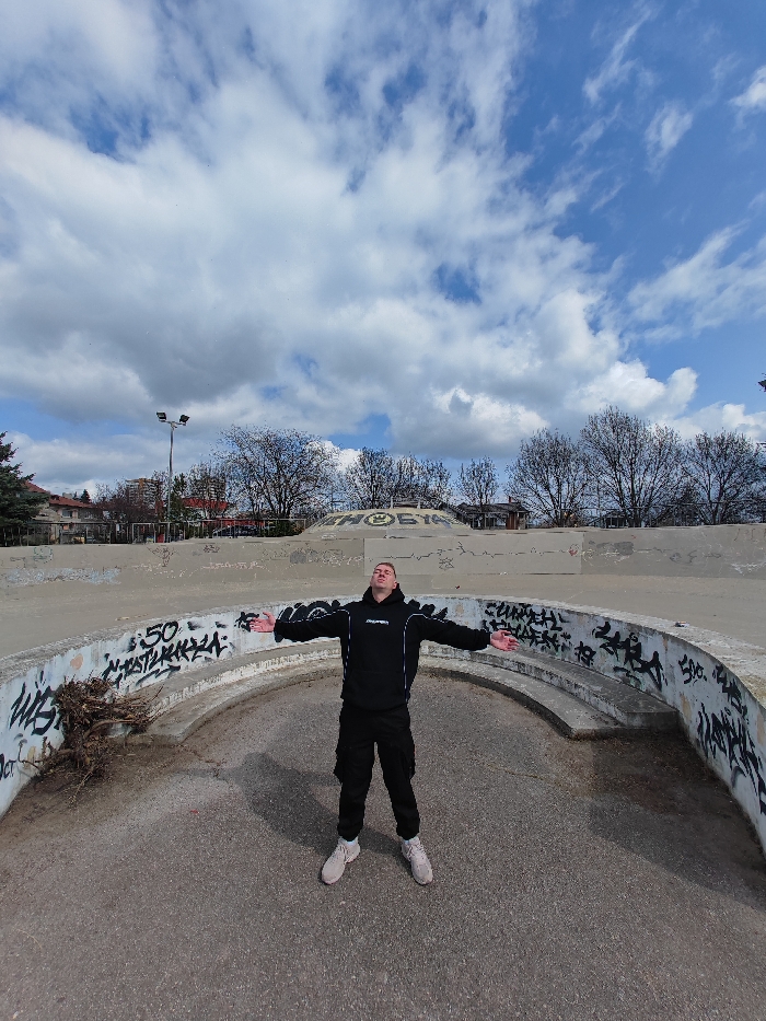 The scene depicts an adult male standing in a concrete skatepark located in Shumen, Bulgaria, under a partly cloudy sky. The subject, a light-skinned male with short hair, is positioned centrally within a curved, graffiti-covered concrete structure in the foreground. He wears a black hooded sweatshirt with white trim and a white logo, black cargo pants, and light-colored sneakers. His arms are outstretched horizontally with palms upward, and his head is tilted slightly skyward, eyes closed or looking up. The curved foreground structure is extensively marked with black graffiti, including the text "50 СТОТИНКИ" visible on its left side, where organic debris is also accumulated. In the background, behind the subject, additional concrete skatepark elements are present, featuring a prominent wall with large black graffiti depicting a clenched fist symbol next to the word "ШОБЯН." Beyond the skatepark, bare deciduous trees, a tall lamppost with two luminaires, and various residential buildings are visible against the sky.