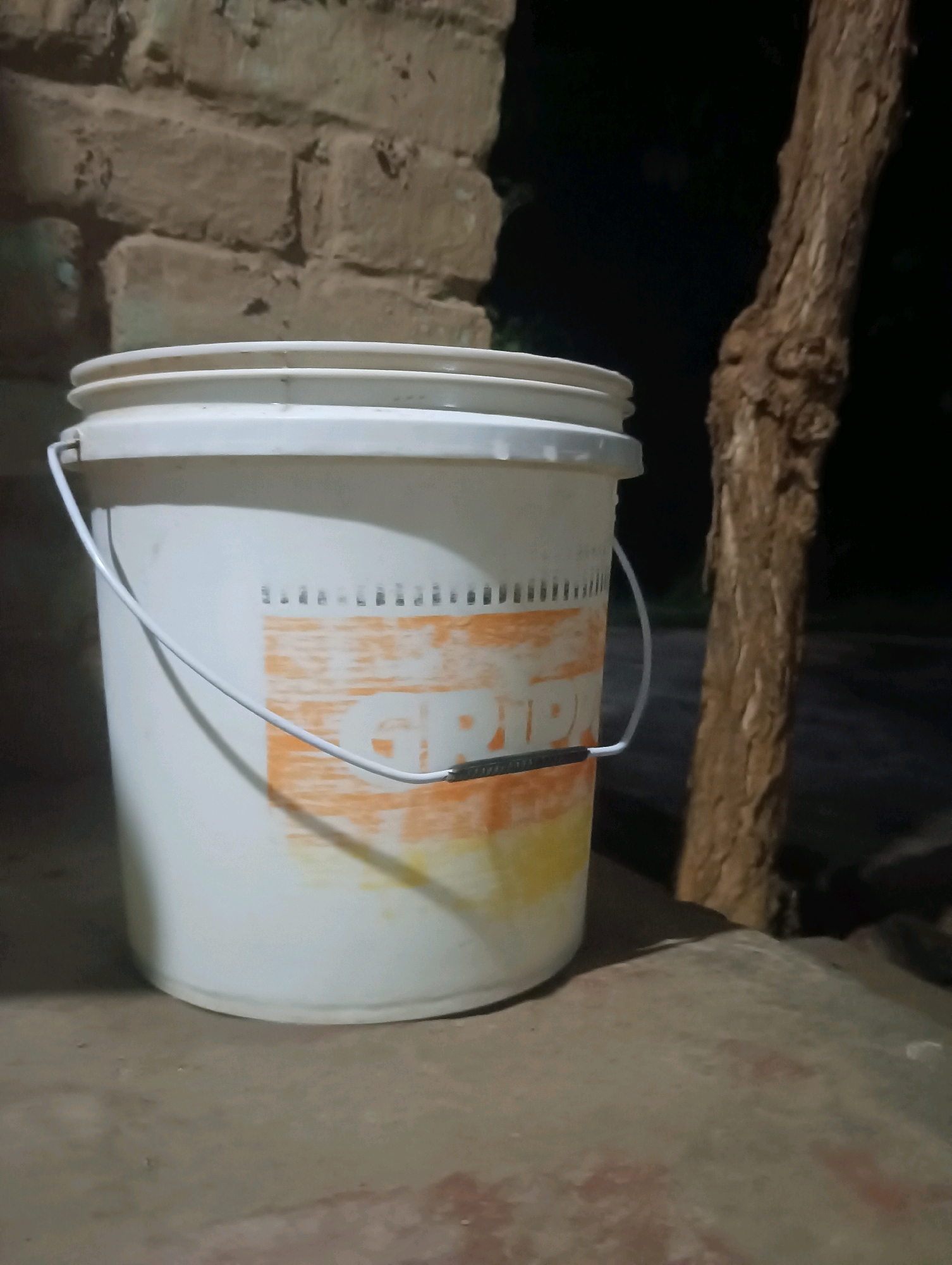 A white plastic bucket with an orange and yellow label is positioned on a textured, light-colored surface. The bucket has a white wire handle attached to its sides. The background consists of a rough, brick wall and a dark, out-of-focus area where a textured tree trunk is visible. The lighting suggests it is nighttime. The scene is located in the city of Kesari, India.