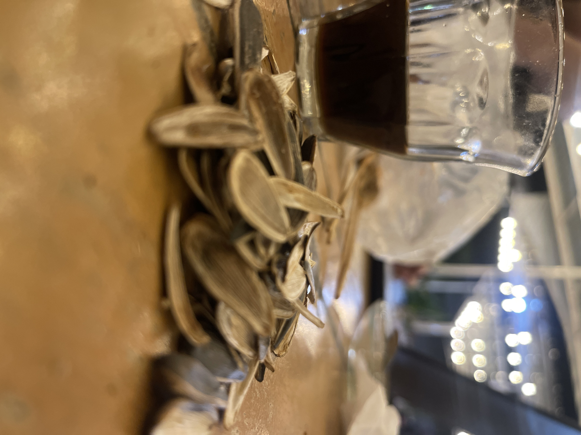 The image shows a close-up of a pile of sunflower seed shells on a wooden table. To the left, a glass of dark liquid, likely coffee, is visible. In the background, out of focus, are blurry lights, suggesting an indoor setting possibly a cafe or restaurant, with a plastic bag and other indistinct objects on the table. The lighting is dim, with the bright background lights creating bokeh effects. The scene appears to be set in Pulokiton, Indonesia, though no specific landmarks or contextual clues confirm this location directly within the image itself.