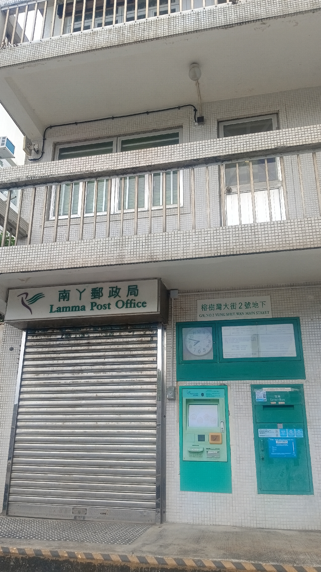 The image shows the exterior of the Lamma Post Office located at G/F, NO.2 YUNG SHUE WAN MAIN STREET, Lamma Island, Hong Kong. The post office has a light-colored signage with Chinese and English text, and an image of a bird. Below the signage is a closed metal roller shutter. To the right of the shutter are two green cabinets mounted on a tiled wall. The left cabinet appears to be an automated postal service machine with a screen and keypad. The right cabinet is a letter box. There is also a clock on the wall to the left of the cabinets. The building appears to be multi-storied with balconies and windows visible on the upper floors. The ground in front of the post office is paved and there are yellow and black striped markings on the edge of the pavement.