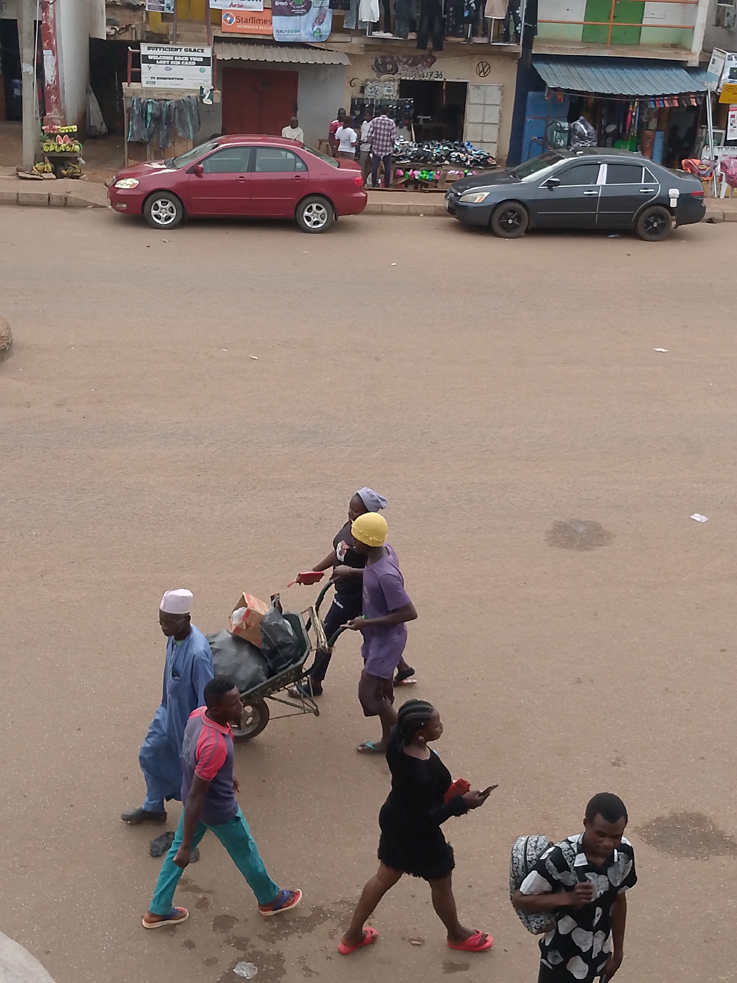 The image depicts a street scene in Bukuru, Nigeria. A red Toyota Corolla and a black Honda Accord are parked on the side of the road. In the foreground, several people are walking, including three individuals pushing a wheelbarrow filled with items covered by a dark bag. A man wearing a white cap and blue robe walks beside the wheelbarrow. Another man, wearing a red and blue shirt with teal pants, is walking in front of them. A woman in a black dress is walking while looking at her phone, and a man in a patterned shirt is walking in the opposite direction carrying a bag. The background shows buildings and shops, with people gathered in front of them. A sign reading "SUFFICIENT GRACE" is visible. The road surface is brownish-grey.