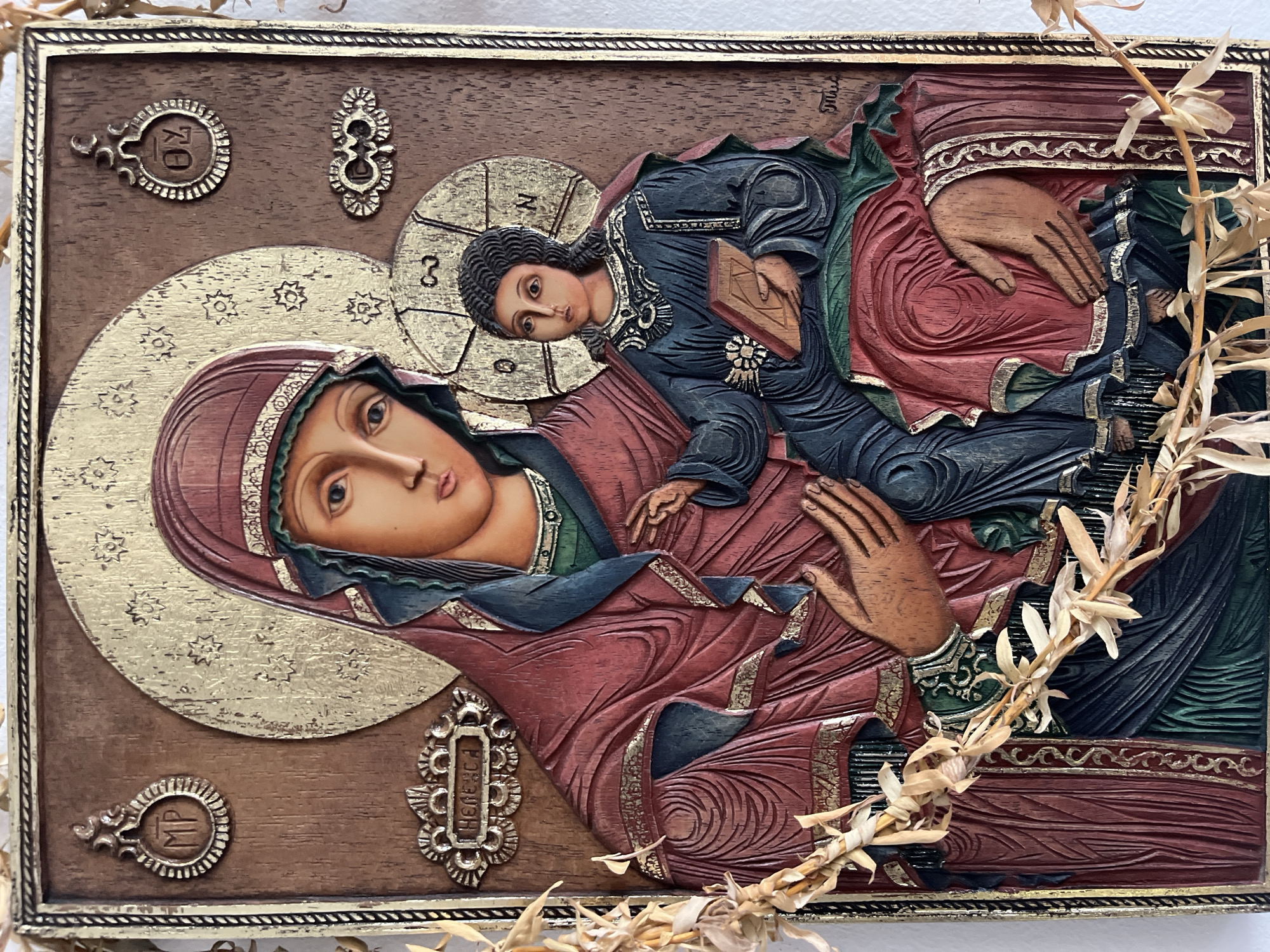 A carved wooden icon depicts the Virgin Mary holding the Christ Child, rendered in relief with painted details and metallic gold-colored accents. The figures are positioned diagonally across the frame. Mary, with a serene expression, wears a deep red maphorion over a dark blue garment. A large, ornate golden halo, decorated with stars, encircles her head, inscribed with the Greek abbreviations "MP" and "ӨУ" (likely "Mother of God").
The Christ Child, with a contemplative gaze, is dressed in dark blue robes with golden trim and holds a closed book in his left hand. His golden, cruciform halo bears the Greek letters "IC", "XC" (for "Jesus Christ"), and "Ο ΩΝ" ("The One Who Is") on its crossbars. His right hand is raised in a gesture of blessing.
The background is dark brown, textured wood. Decorative metallic elements are visible in the corners of the panel. On the bottom right, a small plaque-like element contains the visible Cyrillic/Greek text "НЕВЕСА". Dried, light-colored plant stalks are draped across the right side of the icon. The icon is framed by a decorative border. The overall tone is one of spiritual reverence and traditional iconography.
Location context: Unknown, Unknown