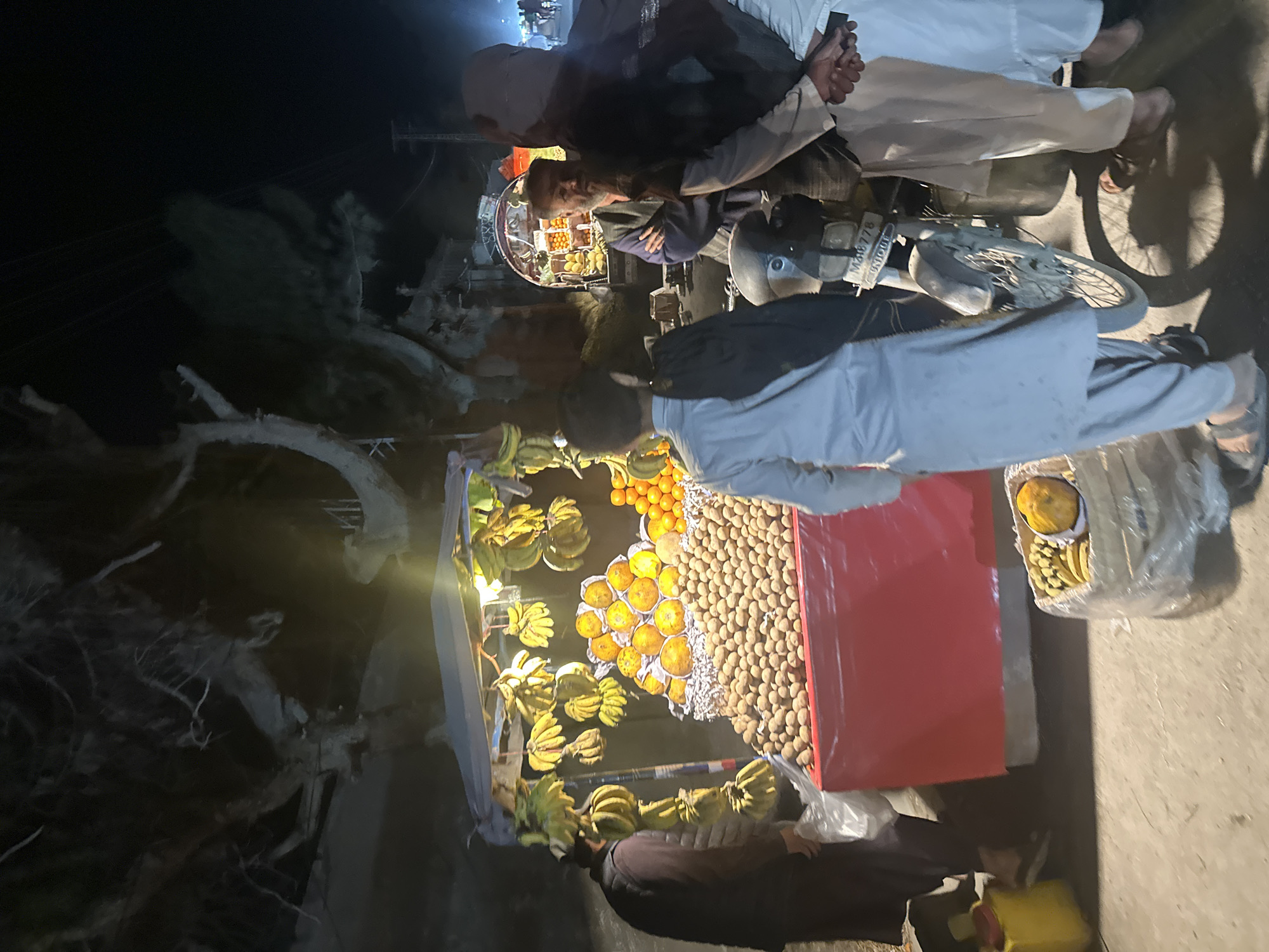 Captured in Quetta City Tehsil, Pakistan, the image shows a dimly lit roadside scene at night. A fruit vendor's stall dominates the foreground, laden with bananas, oranges, and small brown fruits. Several men are present; one is attending to the stall, while others appear to be customers, some standing by a parked motorbike. The environment suggests an urban street with sparse lighting.