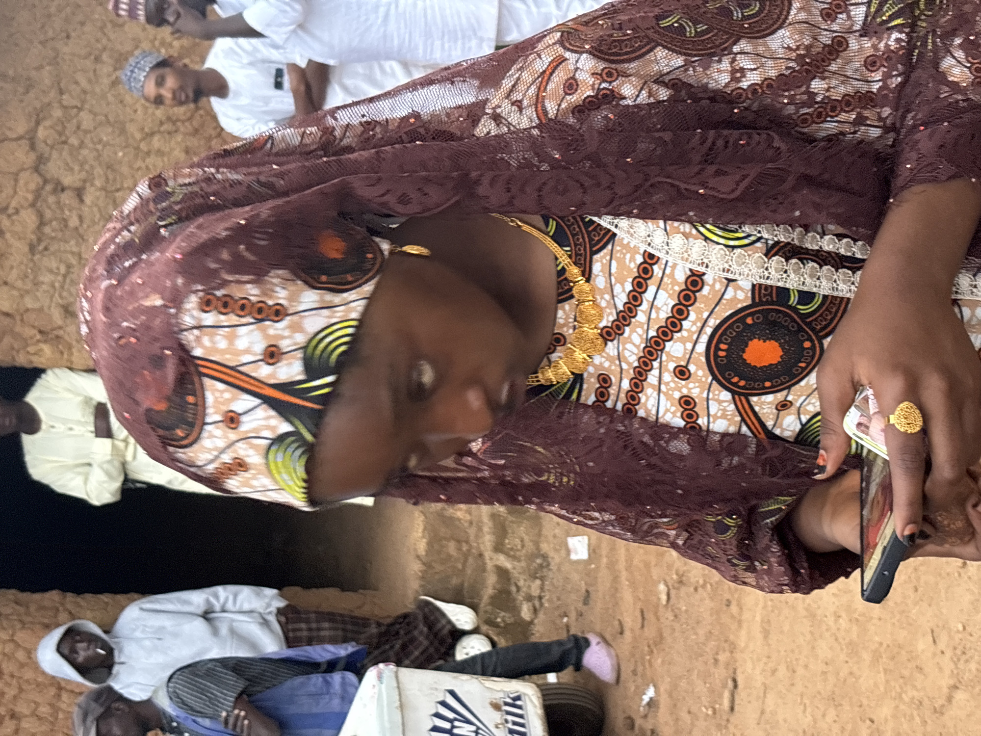 In Jos, Nigeria, a young Black girl, adorned in a patterned headscarf and traditional attire featuring intricate embroidery and lace accents, gazes down at her mobile phone. Her gold necklace and earrings glint in the light, adding a touch of adornment. Her hands, with painted nails, hold the phone, suggesting an act of communication or engagement with the digital world. The background reveals a bustling environment: a man in a white hoodie with a cigarette in his mouth stands in an entryway, while other men in traditional attire are visible further back. A white cooler, possibly for drinks or food, is positioned to the left. The ground is a dusty, reddish-brown earth, typical of the region. The scene depicts a moment of everyday life, blending traditional customs with modern technology in the vibrant city of Jos.