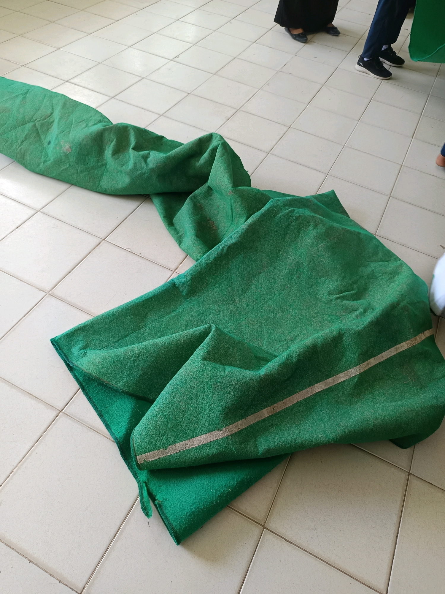 The image shows a green, folded fabric or tarp lying on a tiled floor. The fabric appears to be somewhat worn and has a light-colored stripe running across it. In the background, the legs of several people are visible. One person is wearing dark pants and black athletic shoes, while another is wearing dark shoes and a black skirt or dress. The floor is composed of light-colored square tiles with grout lines. The scene is indoors, as suggested by the tiled floor and the presence of people wearing indoor attire. The specific location in the city of Labuhan, Indonesia, is not discernible from the visual information provided in the image.