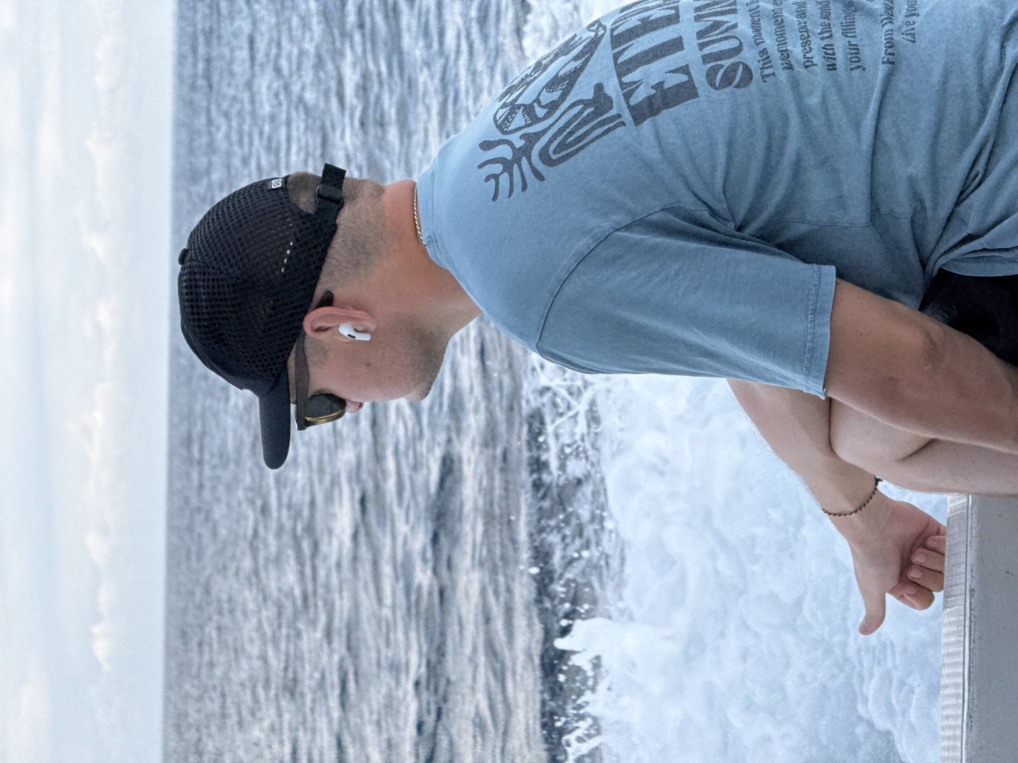 A medium, profile shot of a person on a boat, looking over the side at the churning water. The person is wearing a black baseball cap, sunglasses, a light blue T-shirt with text on the back, and an earbud in the left ear. They are leaning over the side of the boat, with their left hand visible near the edge. The ocean can be seen in the background. The location is near Kampung Toyapakeh, Indonesia.