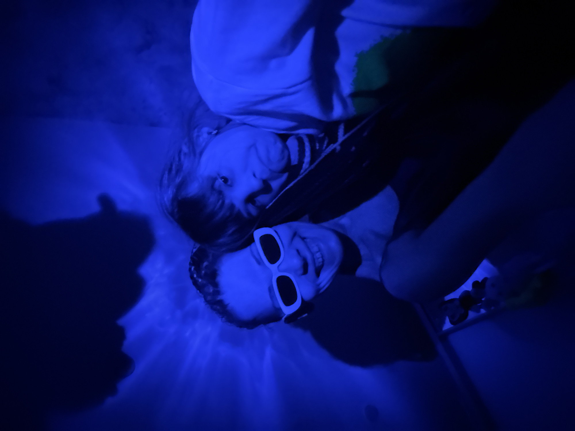 Two individuals are in a blue-lit environment, possibly indoors. One person is wearing white sunglasses and smiling. The other person is wearing a hoodie and looking at the camera. The setting appears to be a bathtub or similar confined space, given the visible light reflections suggesting water or a smooth surface. The city and specific location are unknown.
