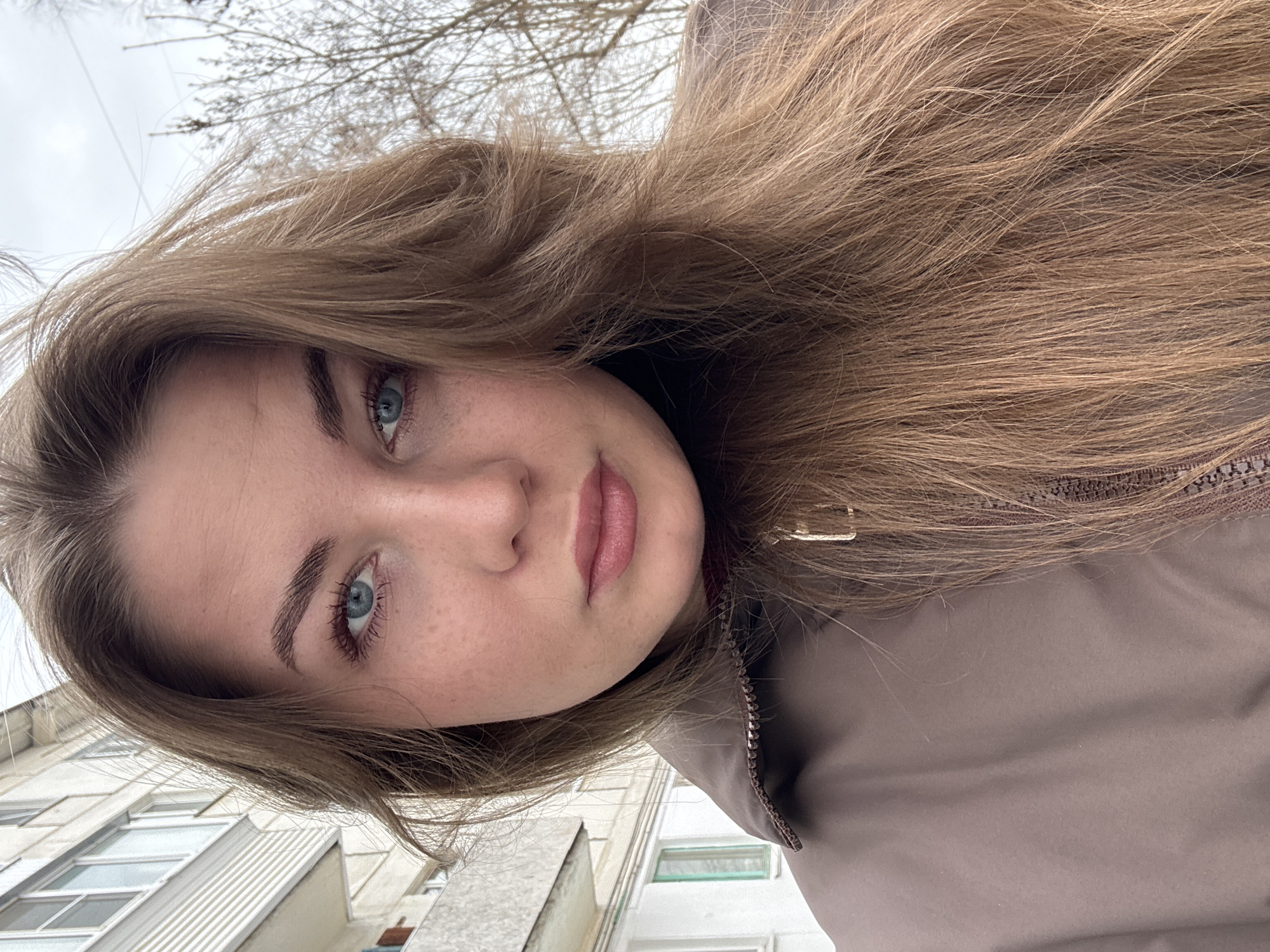 A close-up outdoor portrait captures a young adult female from the chest up, facing directly forward. She has light brown hair, blue eyes, fair skin with visible freckling, and her lips are slightly parted. She wears a brown or taupe collared jacket with a visible metallic zipper pull, over a darker garment. The background on the left features a multi-story, light-colored residential building with rectangular windows and a visible balcony. The background on the right shows bare tree branches against an overcast sky. The scene is situated in the city of Chaikovsky, Russia.