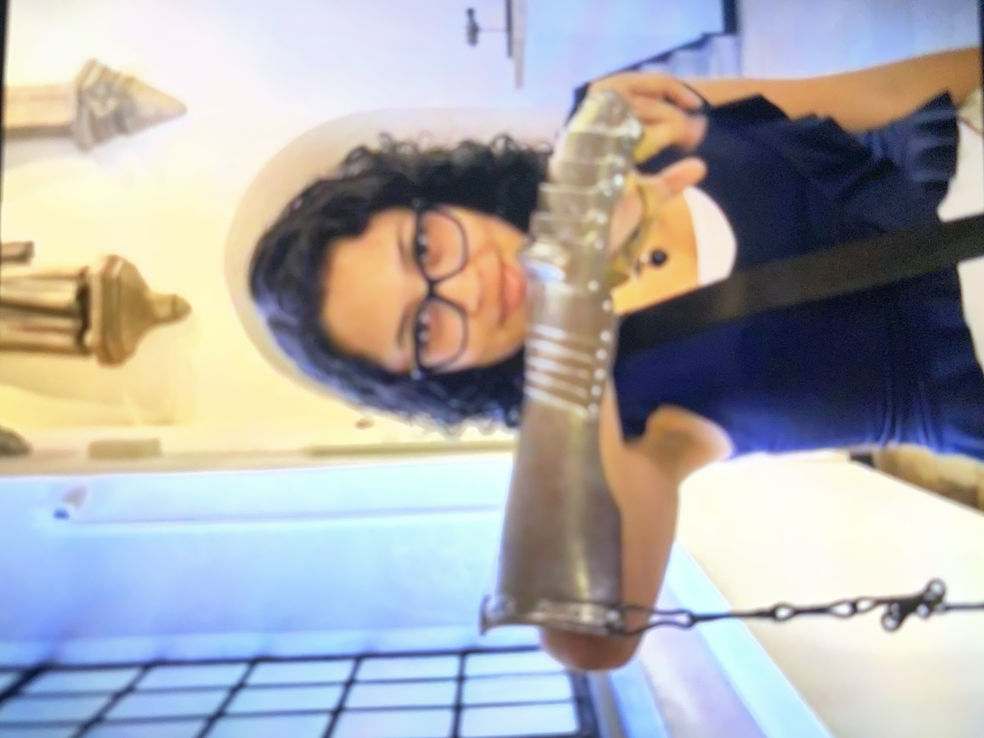 A woman with dark curly hair and glasses is holding a medieval gauntlet. She is wearing a dark blue dress with a white collar. Behind her, on the wall, are some ornate golden objects. In the foreground is what appears to be a metal grate.