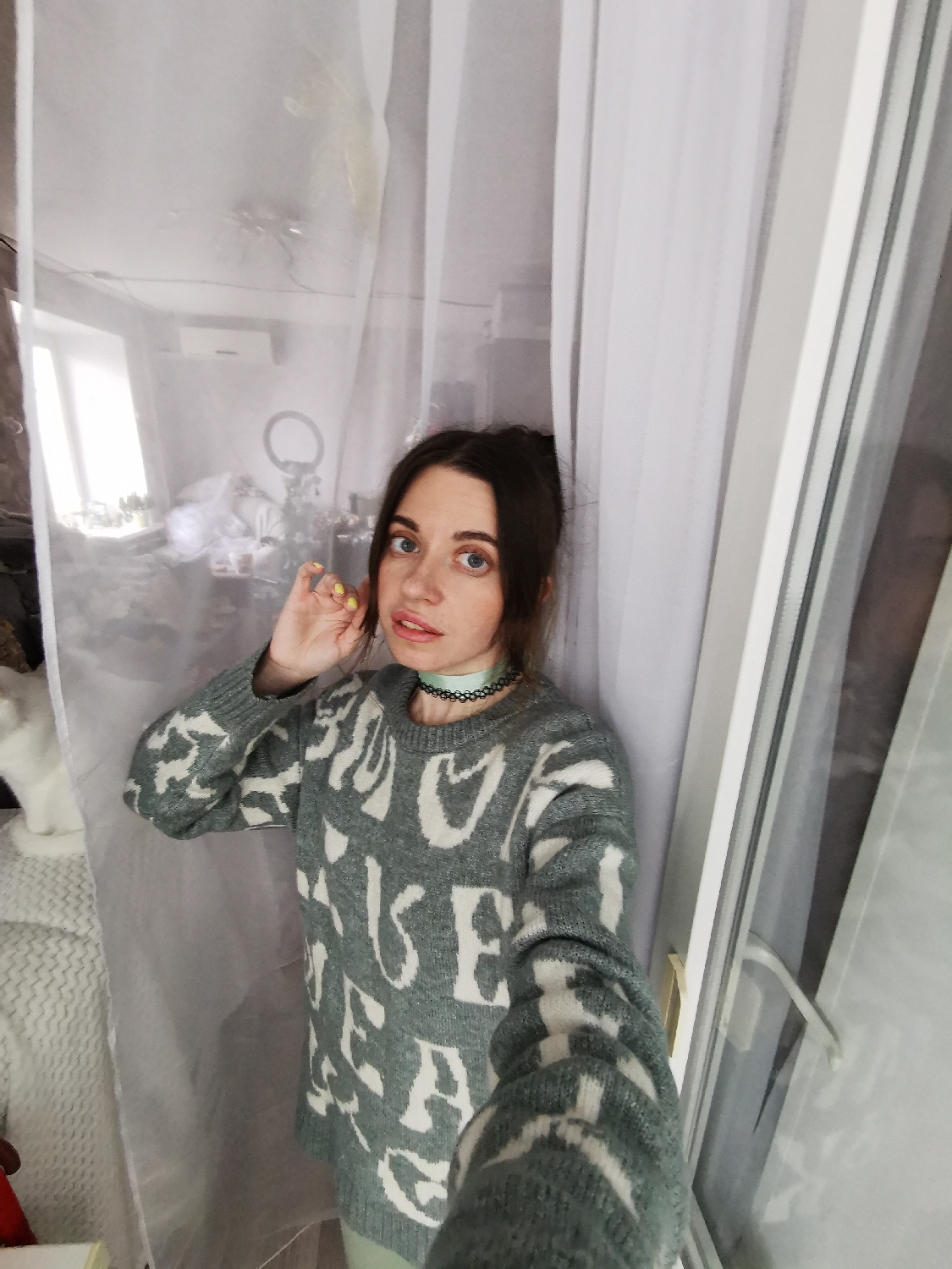 A young woman is depicted in a medium shot, looking directly at the camera with wide blue eyes and a slightly pouting expression. Her dark hair appears to be pulled back, and she is wearing a greyish-green knitted sweater adorned with large, abstract white shapes that resemble jumbled, overlapping letters. A light green choker is visible around her neck, and she has yellow nail polish on her fingers. Her right hand is raised near her face, with her fingers lightly touching her hair or ear.
She is standing indoors, positioned between sheer white curtains that appear to frame a large window or glass door. To her left, a white cat is partially visible on a textured white surface, possibly a blanket or piece of furniture.
The background behind the woman is a reflection visible through the glass, showing an interior room. This reflection includes a window, an air conditioner unit mounted on a wall, and a prominent ring light, suggesting a home or studio setting. The lighting in the image indicates it is daytime, with ample natural light illuminating the scene. The white shapes on her sweater are decorative and do not form any discernible, coherent words or phrases.