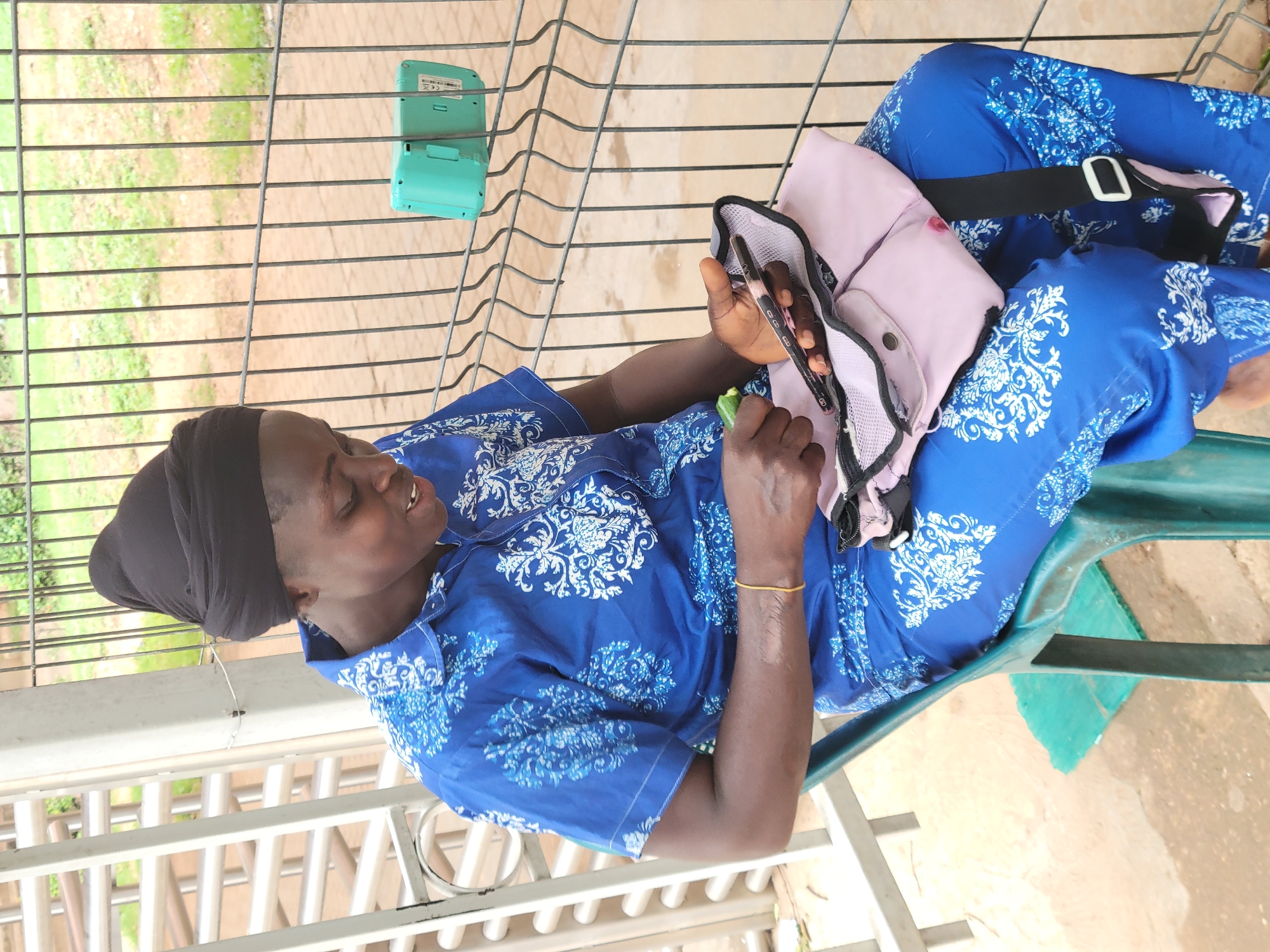 An African woman wearing a blue patterned garment and a black head wrap sits on a green plastic chair in an outdoor setting. The woman is looking down at a smartphone held in her hand. A pink bag sits on her lap next to the phone. A teal-colored device is affixed to the metal railing behind her. The scene appears to be on a patio or veranda with a view of vegetation through the railing. The location is Abuja, Nigeria.
