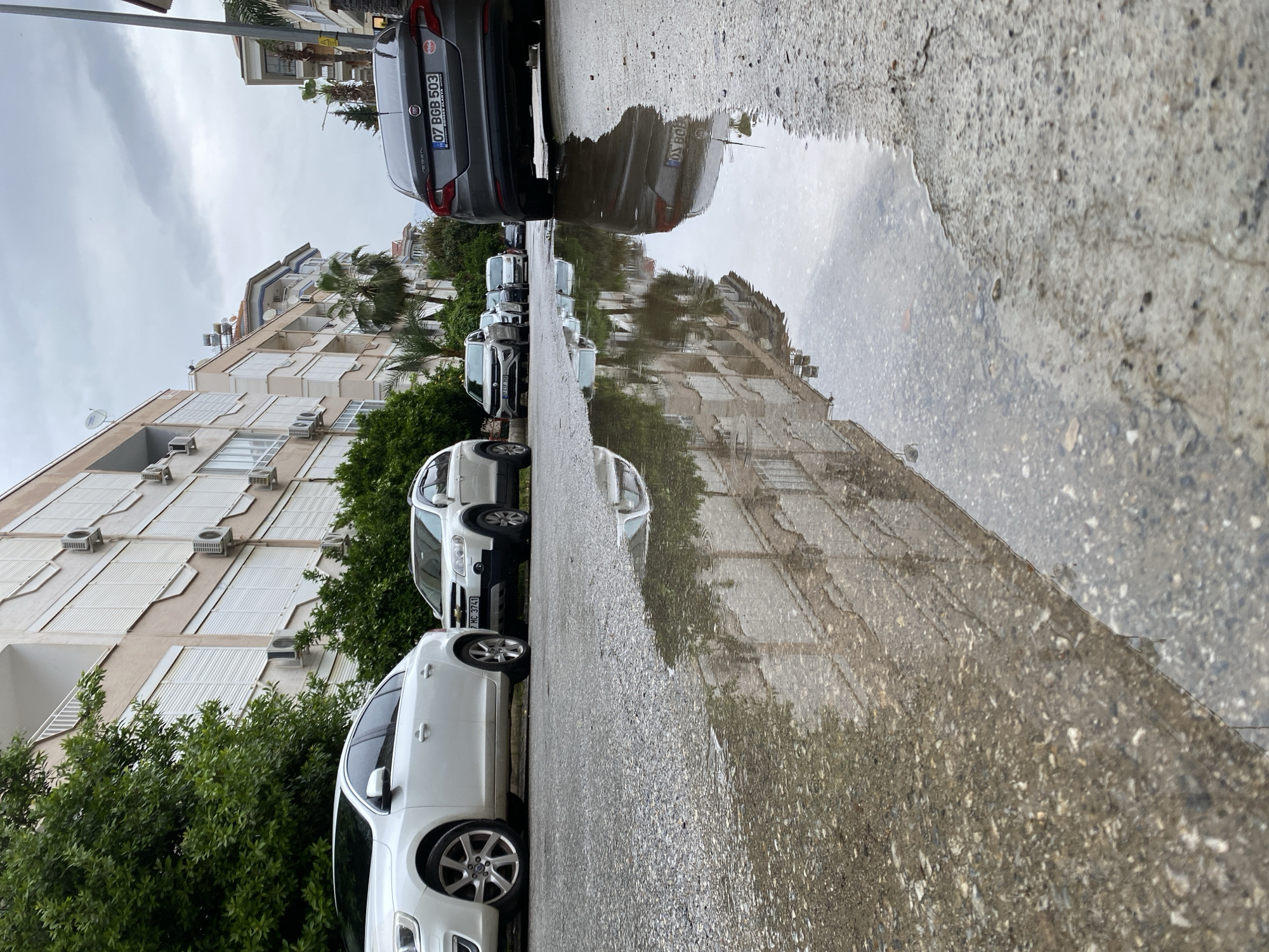 The image shows a street scene in Alanya, Turkey on a cloudy day after a rainfall. A puddle on the asphalt reflects the buildings and cars parked along the street. The buildings appear to be residential apartments, with multiple stories and numerous air conditioning units visible on their facades. Several cars of various makes and models are parked parallel to the curb, with license plates visible on some. The trees and foliage add some green to the scene, contributing to the neighborhood's aesthetic. The overall impression is of a typical residential street in an urban setting.
