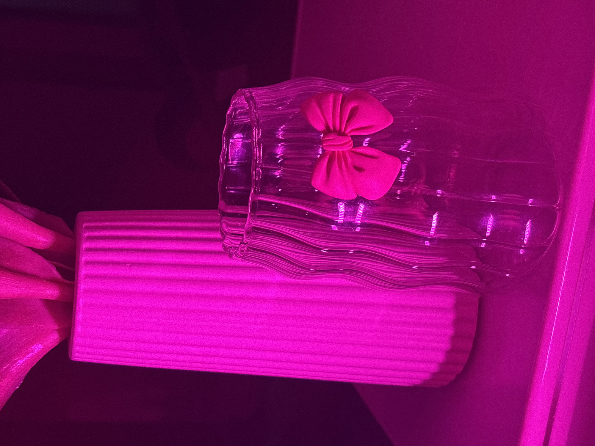 The image depicts a still life arrangement with two vases on a surface, bathed in intense pink lighting. On the left is a tall, ribbed, pink vase containing what appears to be foliage. To its right is a shorter, clear glass vase with a wavy texture. Attached to the front of the glass vase is a bright pink bow. The background is dark and indistinct. There is no discernible context related to the city of Мещёры, Russia, or any people or actions.