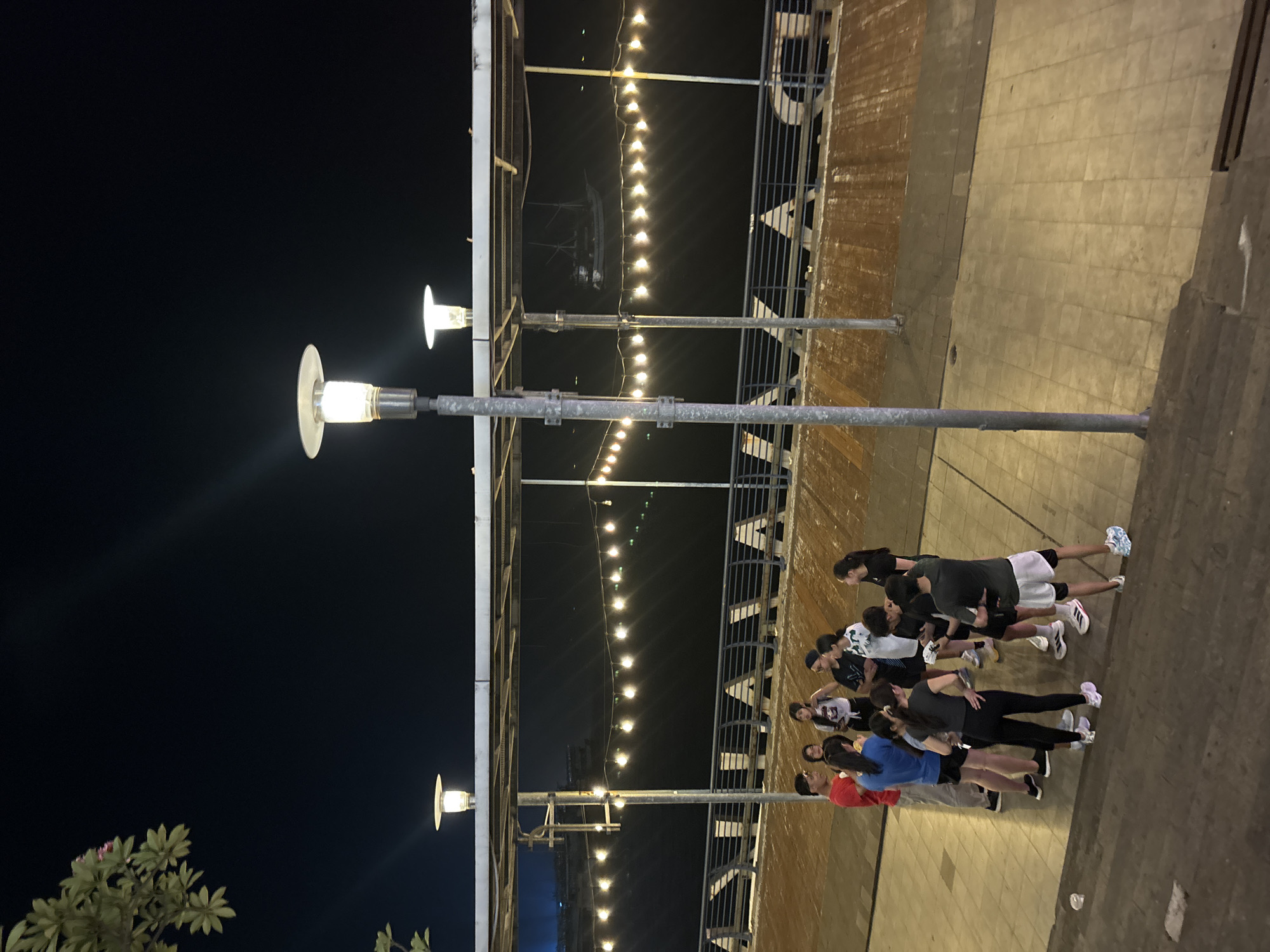 A group of approximately ten young adults, dressed in athletic wear, are gathered on a paved walkway at night. They are standing in a huddle, appearing to be engaged in conversation or activity. The walkway is adjacent to a body of water, possibly a river or canal, with a railing and decorative lights strung overhead. In the background, there are distant city lights and a silhouette of a large boat. The scene suggests an outdoor gathering or event in an urban waterfront area.