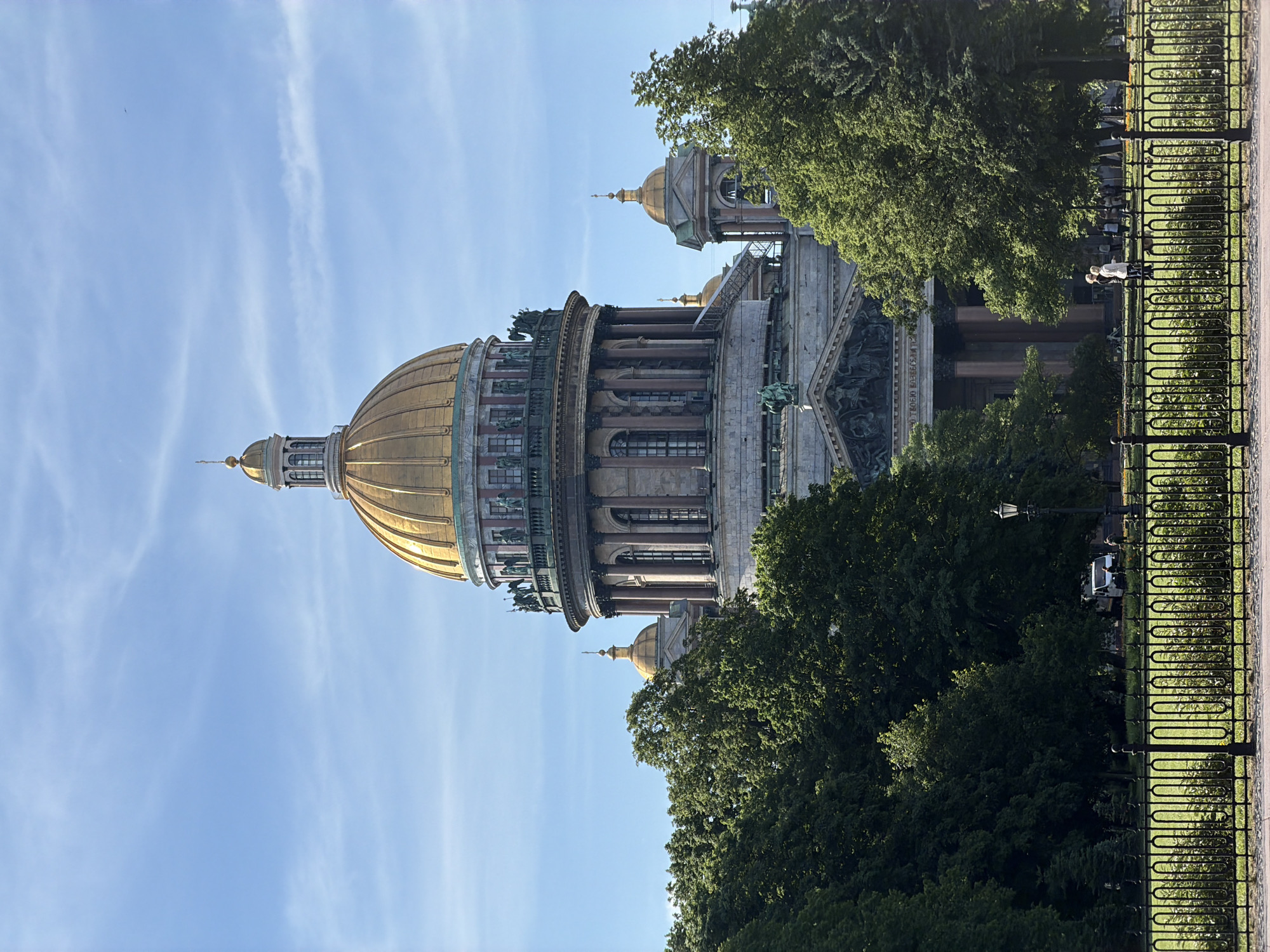 The image captures a clear, sunny day with a bright blue sky featuring wispy white clouds. Dominating the center is the grand dome of St. Isaac's Cathedral in Saint Petersburg, Russia, gilded and prominent against the sky. The cathedral's stone facade, portico, and smaller golden domes are visible, partially obscured by lush green trees in the foreground and middle ground. A black wrought-iron fence runs across the bottom of the image, separating a well-maintained green lawn from a pathway. Several people are visible walking on the path behind the fence, and a white van is parked in the distance. The overall scene depicts a peaceful day in a park setting with the iconic cathedral as the backdrop.