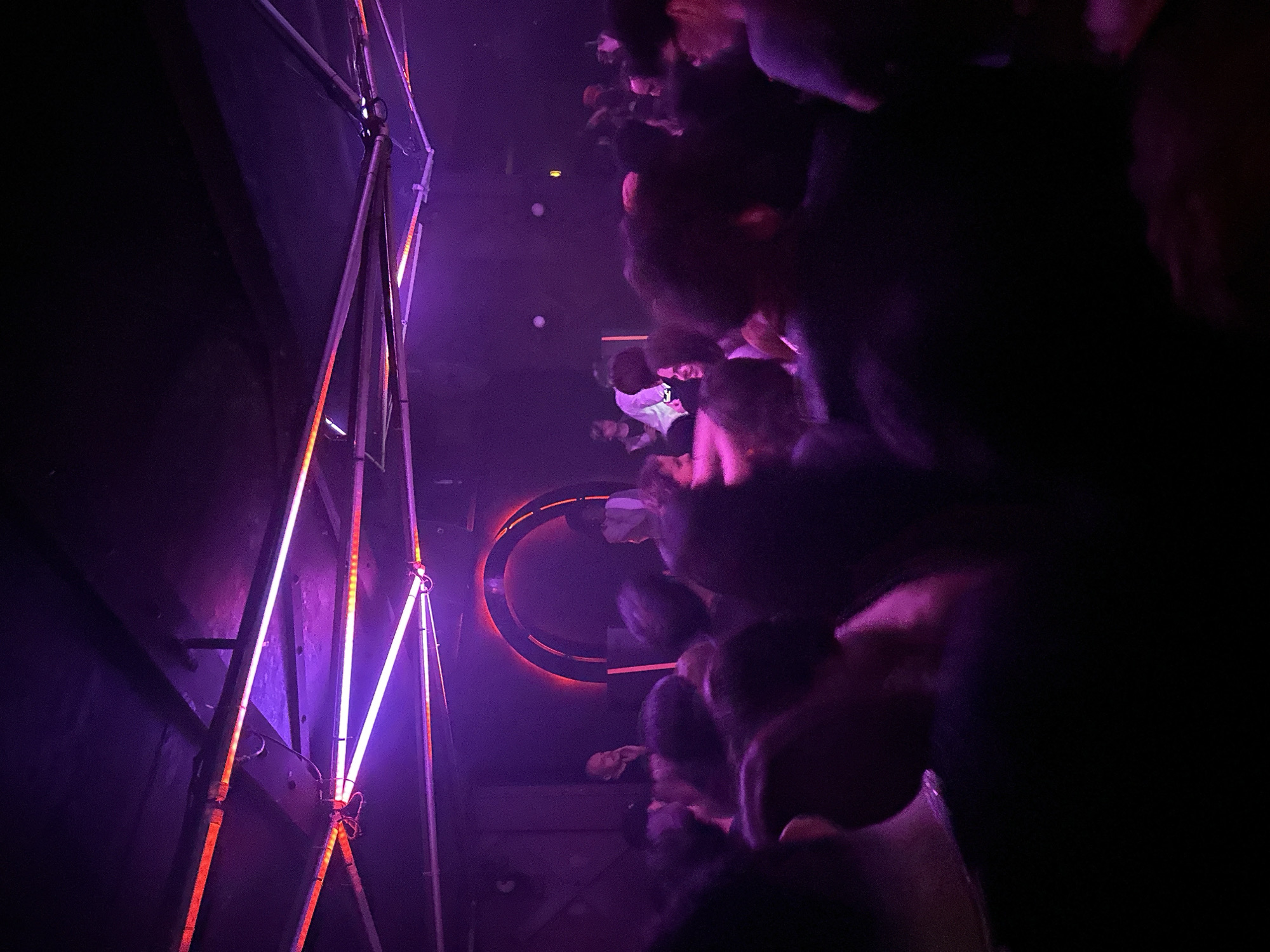 This image depicts a crowded, dimly lit indoor space, identified as a nightclub or music venue in Paris, France. The environment is illuminated predominantly by artificial purple and red light sources. Numerous linear light tubes, emitting both purple and red light, are visible affixed to the dark ceiling, which displays exposed structural elements. In the background, a large, glowing red circular arch serves as a prominent focal point.
A dense crowd of individuals occupies the majority of the frame, their figures largely silhouetted against the ambient lighting. People are standing, facing generally towards the background area. In the background, behind the main crowd, a bearded man is partially visible to the left of the red arch, observing the scene. Another individual is partially visible within or near the glowing arch, and further to the right, other patrons or staff are present, including one person wearing a lighter-colored garment. The overall scene suggests a lively social gathering within an entertainment setting.
