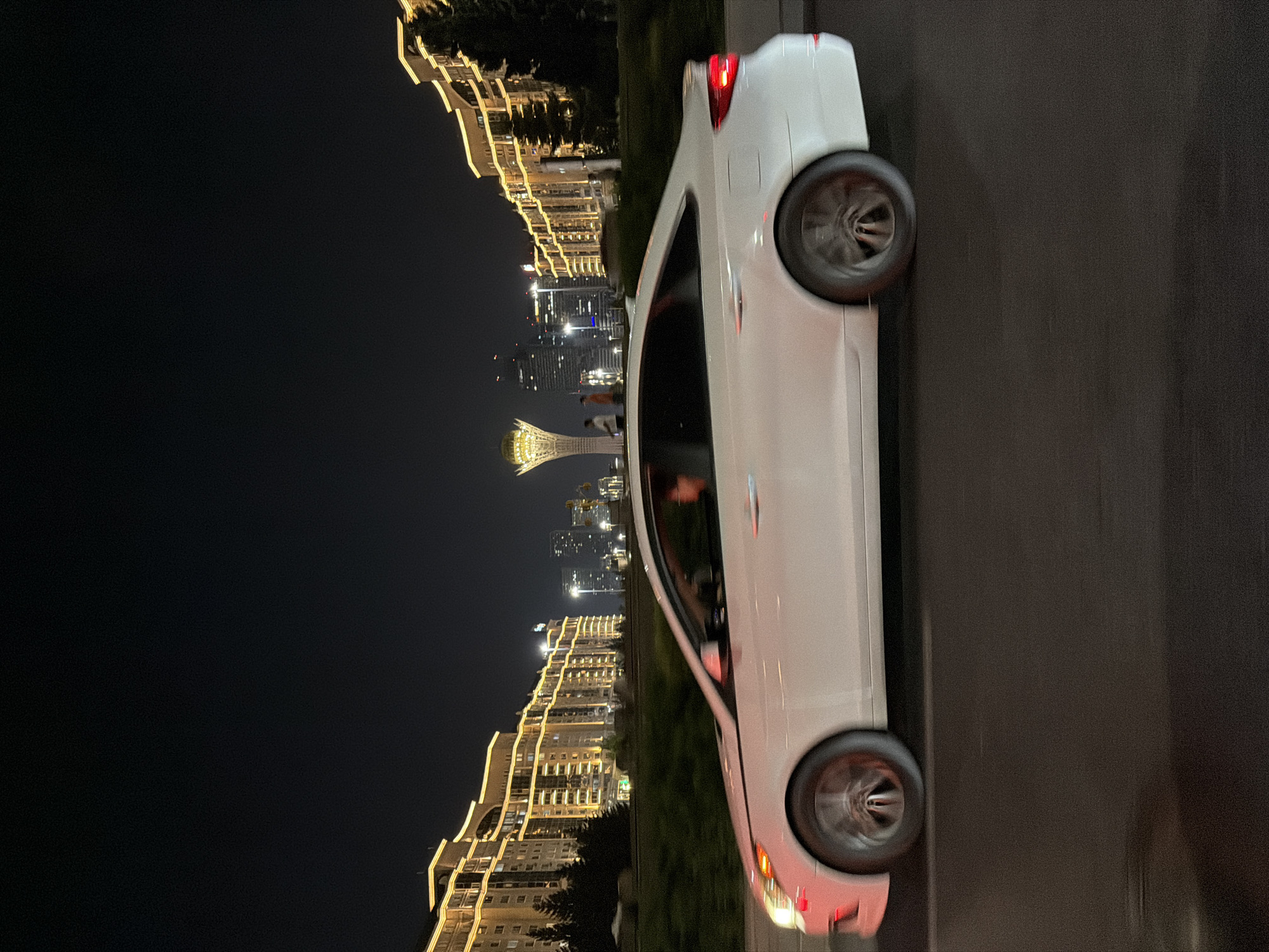 A white sedan is captured in motion at night on a city street, possibly in Astana, Kazakhstan, as evidenced by the distinctive architecture. The car is a blur of motion, suggesting a fast shutter speed. The driver, a person with dark hair, is visible through the side window. The car's headlights and taillights are illuminated. In the background, illuminated buildings line both sides of the street, displaying a pattern of yellow lights. A prominent, dome-like structure with lights is visible in the distance, a common architectural feature in Astana. Two figures are seen standing near this structure. The overall scene is dark, characteristic of nighttime, with artificial lighting dominating the visual landscape.