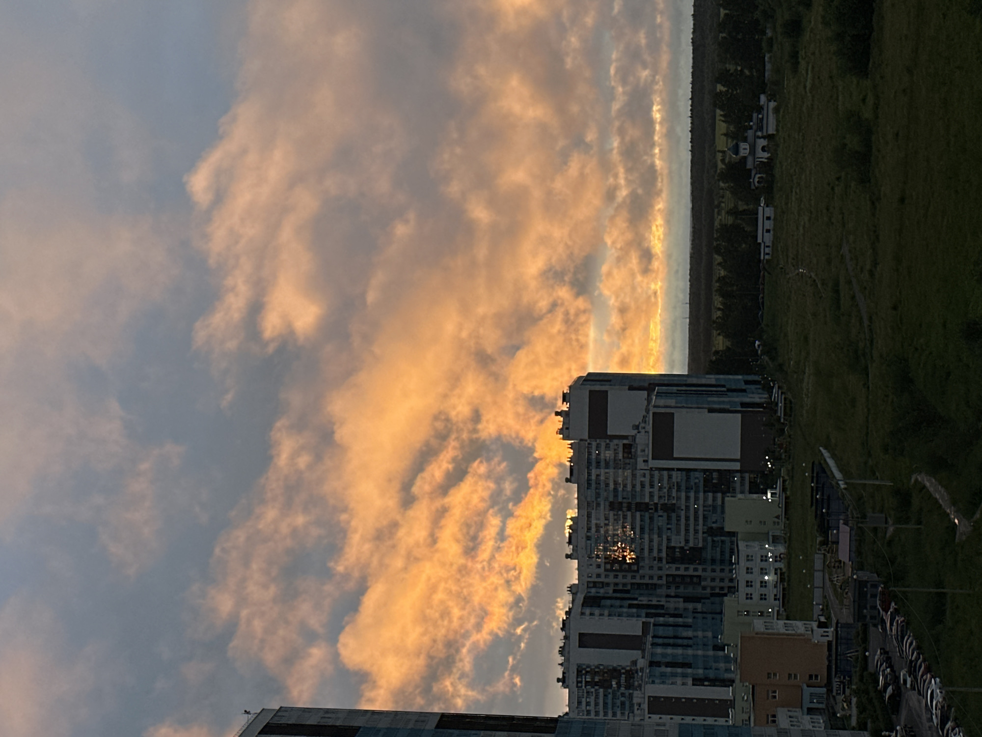 This media file depicts a city skyline at sunset, with a prominent display of orange and yellow clouds against a pale blue sky. The scene is dominated by several tall, modern residential buildings with an array of windows and balconies, reflecting the warm hues of the setting sun. In the foreground and on the right side of the frame, the sky takes up a significant portion of the image, showcasing the dramatic cloud formations.
Below the sky and buildings, a strip of dark green trees and a grassy field can be seen, suggesting a suburban or exurban setting. Further to the right, a row of parked cars is visible, indicating a residential area. The overall impression is one of urban development encroaching on a natural landscape. The time of day is clearly sunset, given the vibrant colors in the sky and the dimming light. The weather appears to be clear with scattered clouds. There is no visible text in the image. The mood is peaceful and contemplative, with the beauty of nature juxtaposed against the man-made structures.