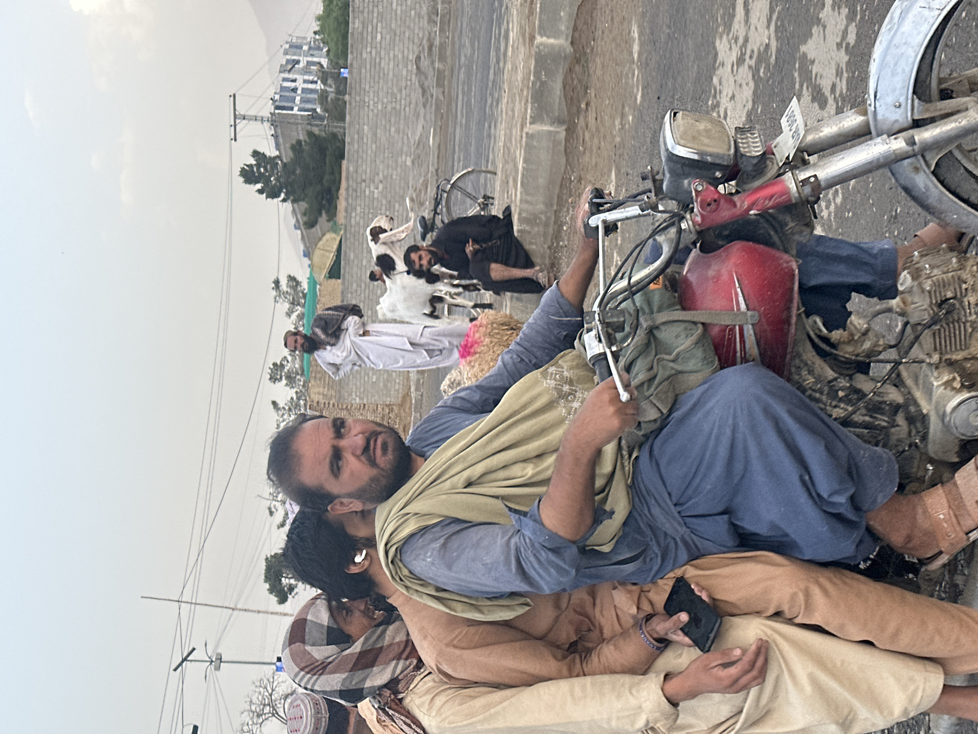 The image depicts three men on a motorcycle near what appears to be a roadside gathering in Quetta City Tehsil, Pakistan. The man in the middle, wearing a blue shirt and beige shawl, is seated on the motorcycle and looking at the camera. The man behind him is holding a black smartphone. Other people are seen further back on the side of the road. Buildings are visible in the background.